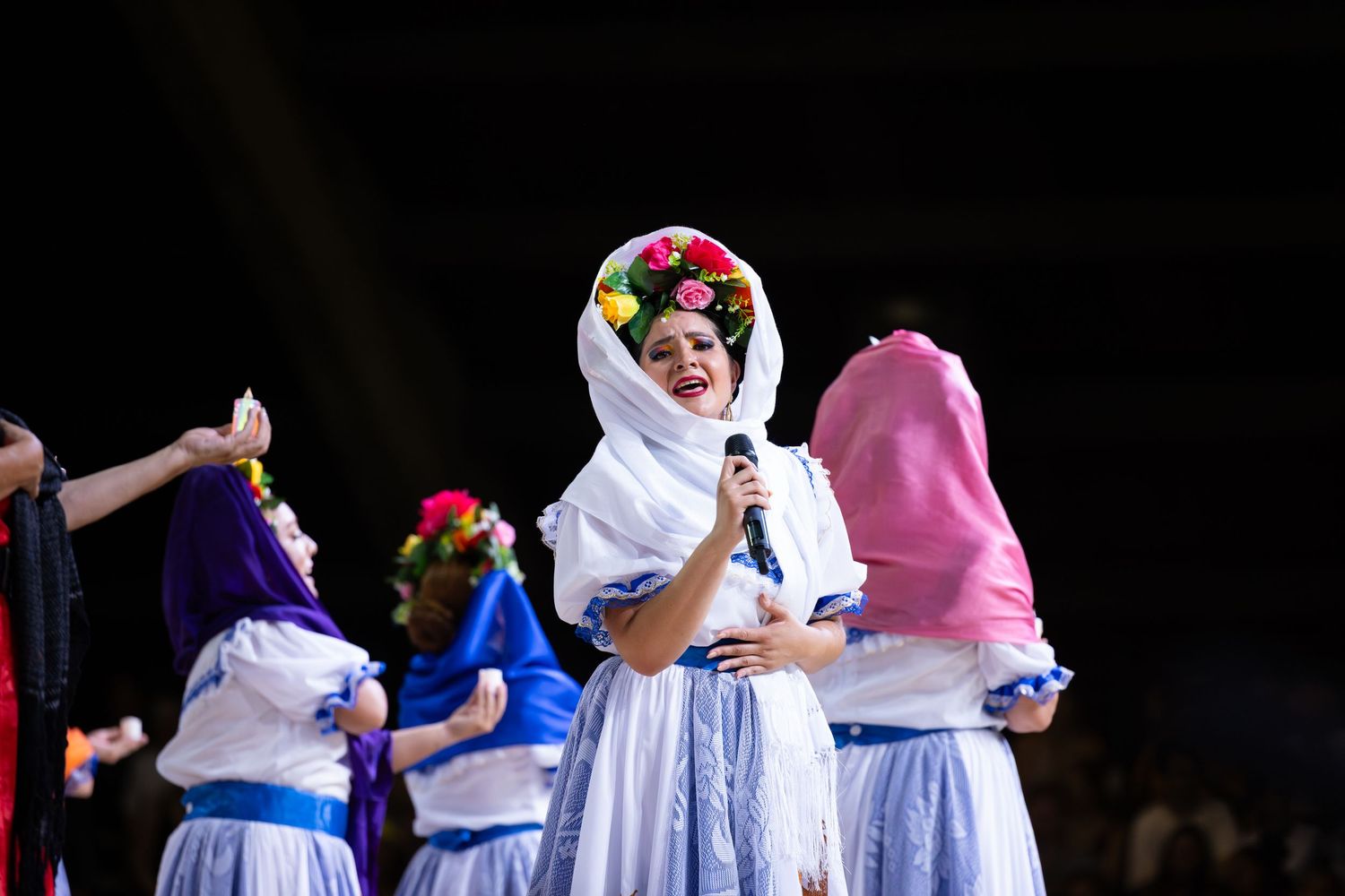 Traditional folk dancers in white dresses and blue sashes perform a synchronized routine on a dark stage.