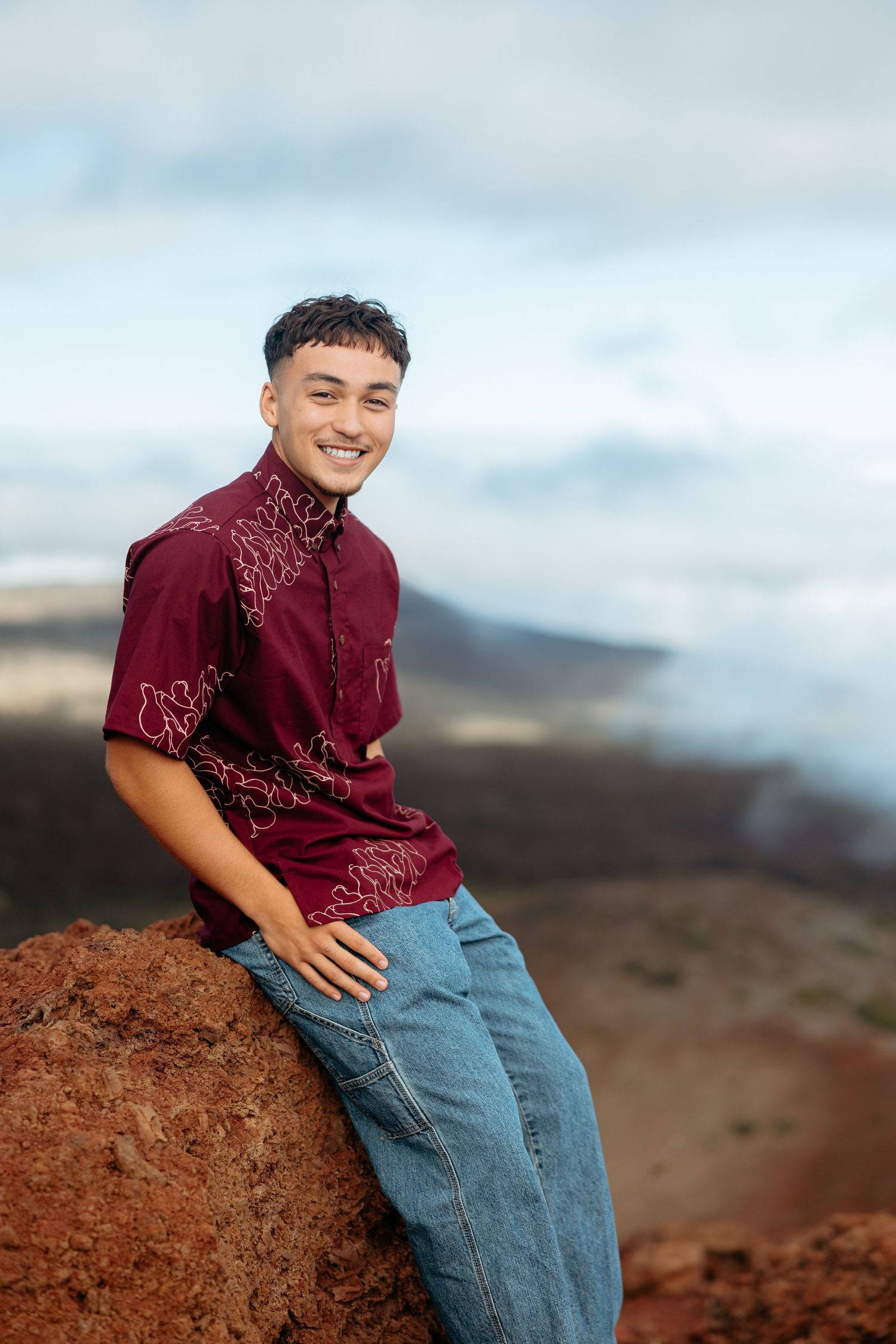 Person sitting on red volcanic rocks in casual attire against a cloudy mountain backdrop.
