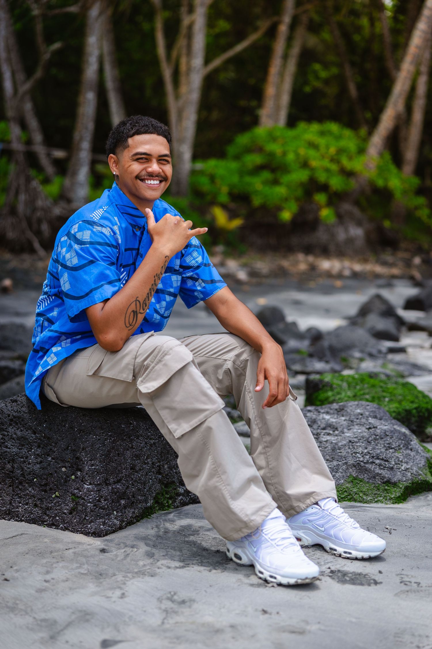 Young man in blue floral shirt and cargo pants sits on rocks, smiling and giving shaka sign with tropical trees behind.