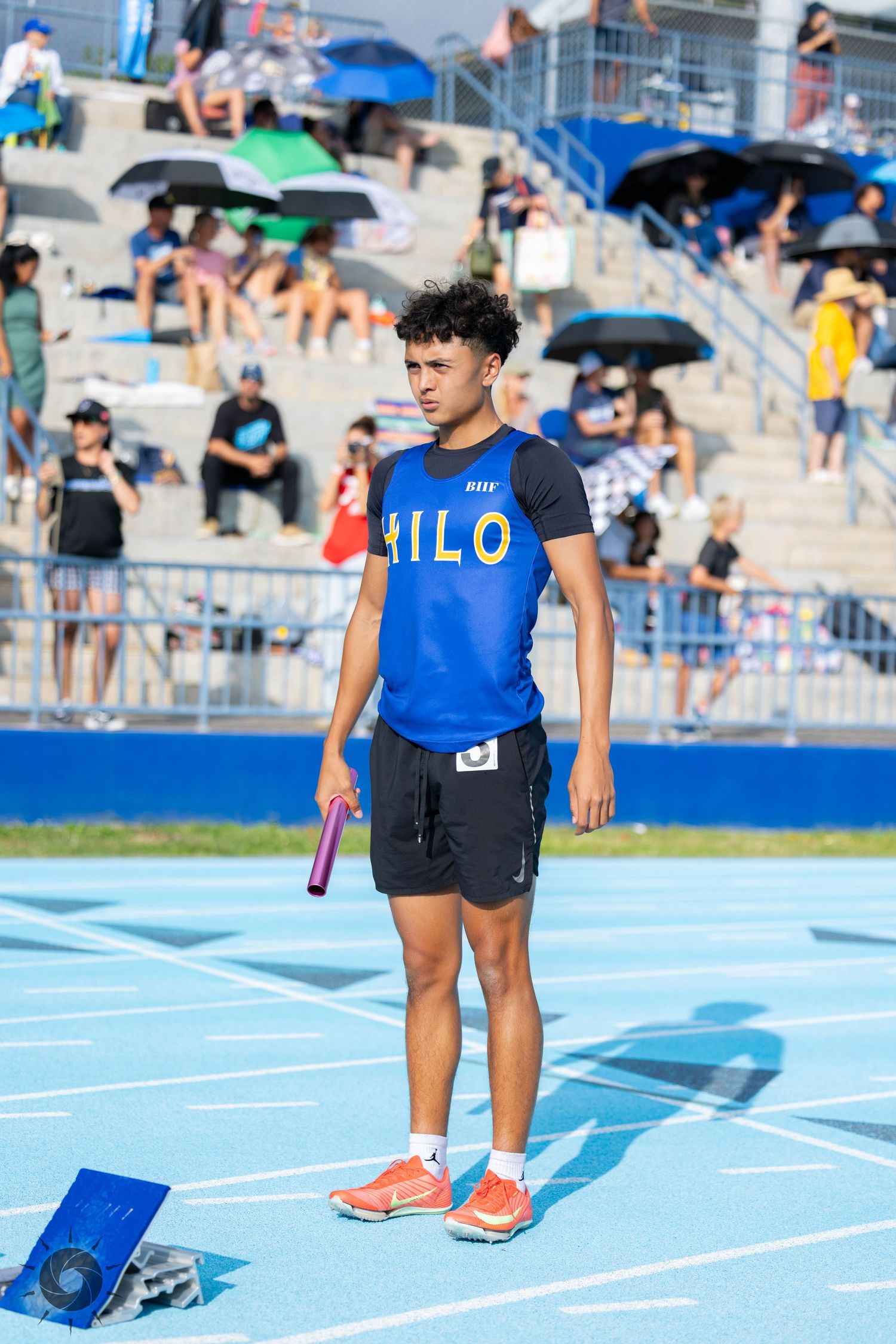 Athlete in blue uniform standing on a bright blue running track with sports equipment nearby.
