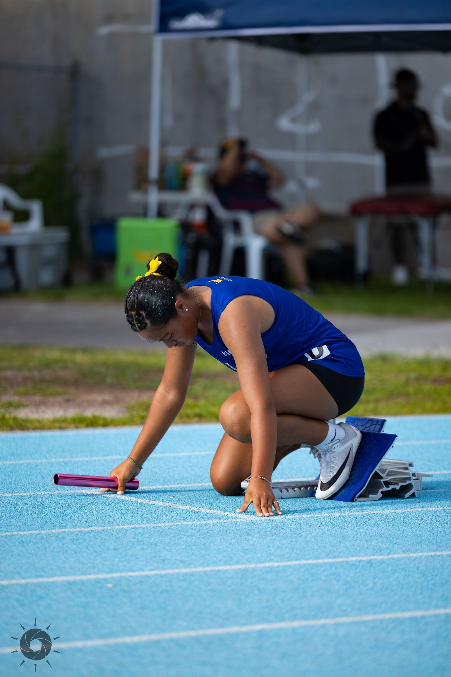 Track athlete in blue uniform crouches at starting position on blue running track preparing for race start.