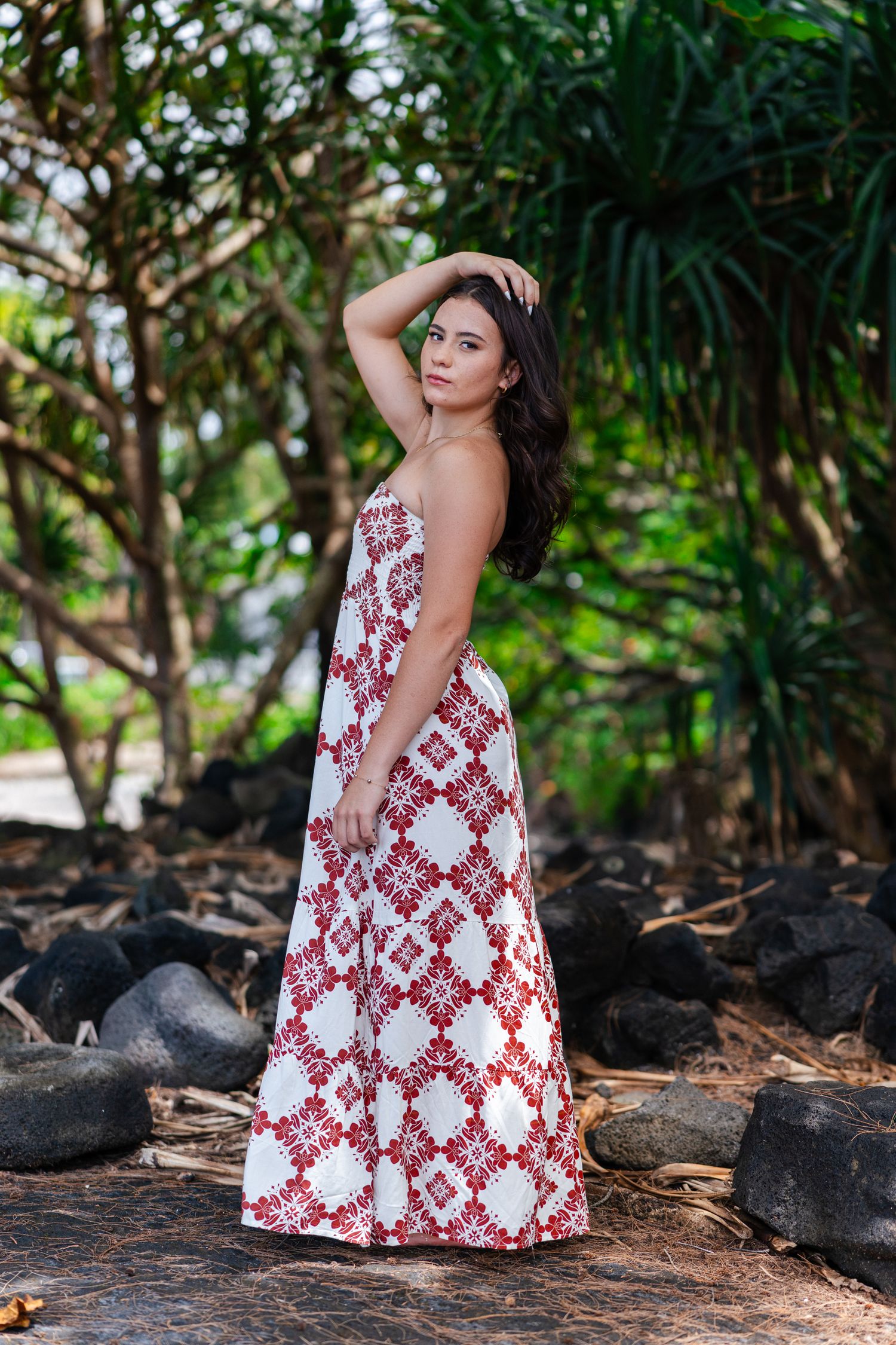 Woman in red and white floral maxi dress posing among tropical trees and volcanic rocks.