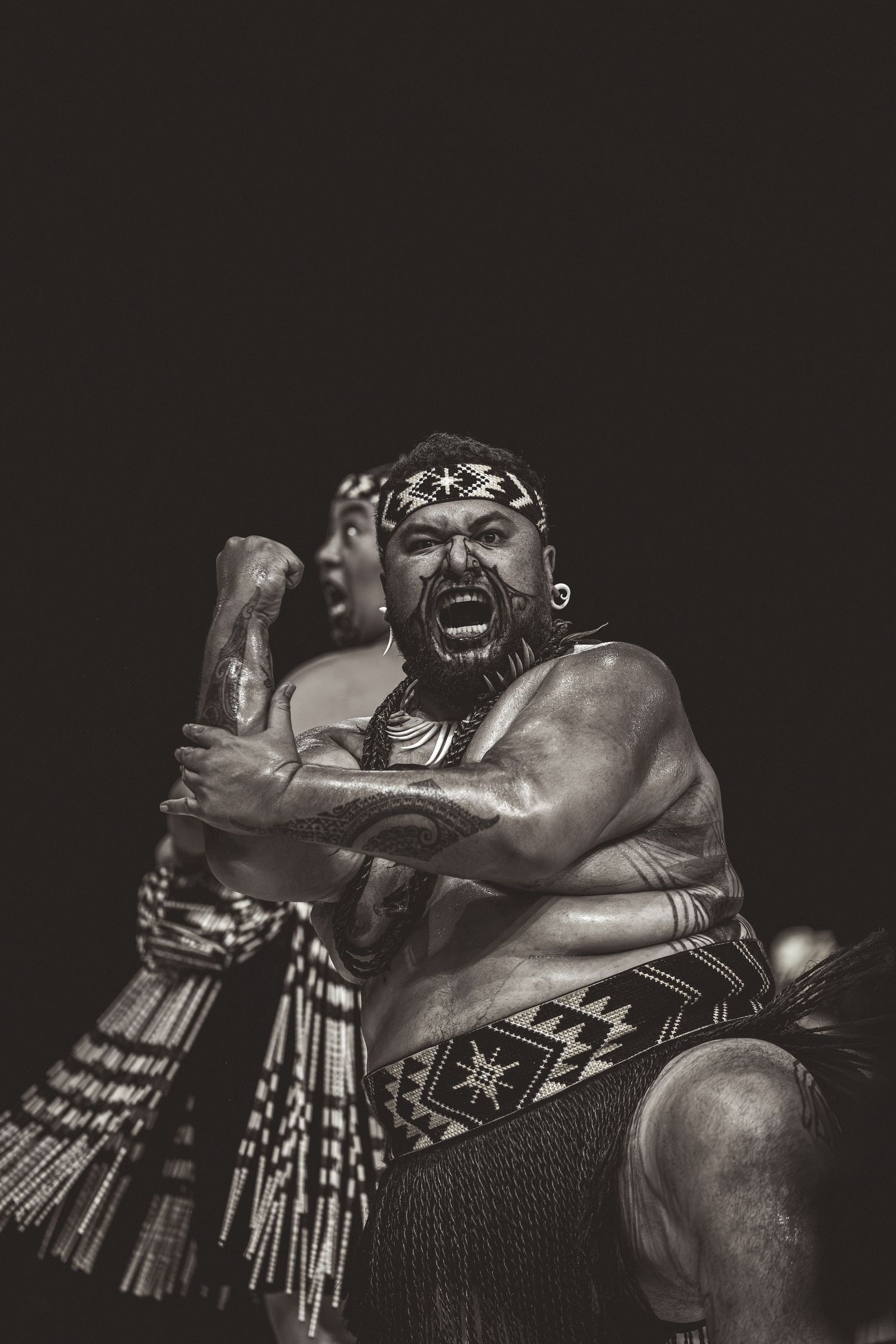 Dramatic black and white photo of a Maori warrior performing traditional haka dance with intense facial expression.