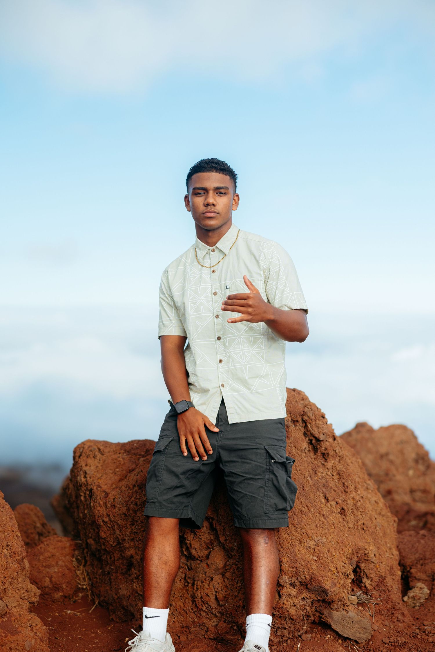 A person in a white shirt and dark shorts sits on red volcanic rocks against a blue sky background.