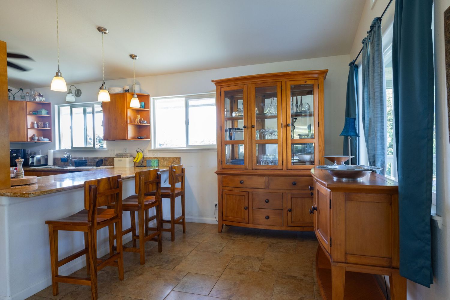 Bright kitchen interior with wooden cabinets, breakfast bar, and teal curtains features tile flooring and natural lighting.