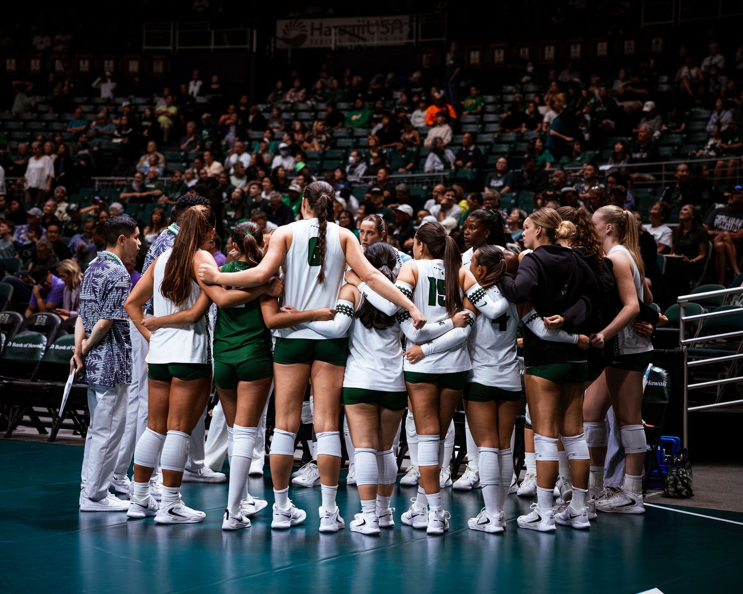 College volleyball team huddles together on court in green and white uniforms during a game at an indoor arena.
