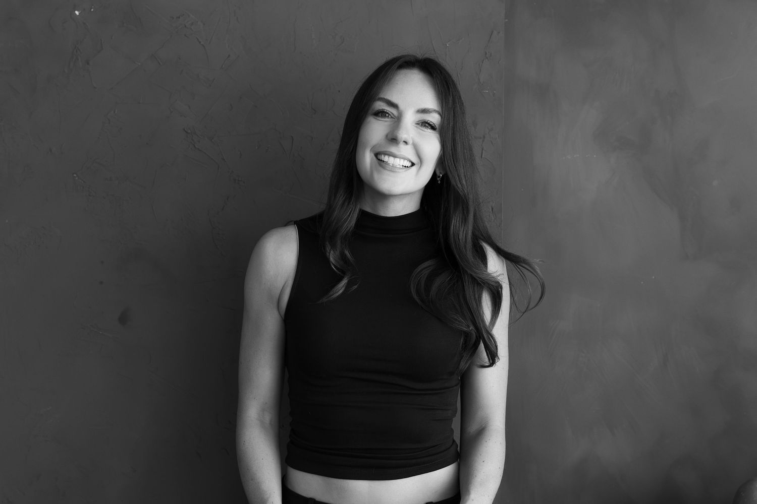 Photographer Caro standing in front of a wall and smiling into the camera in black and white
