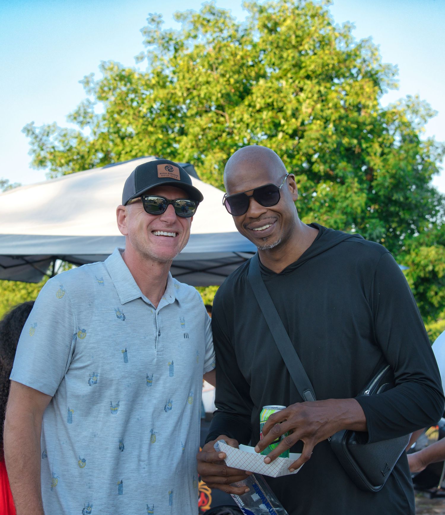 Two men, one wearing a gray shirt and the other a black shirt, stand together outdoors.