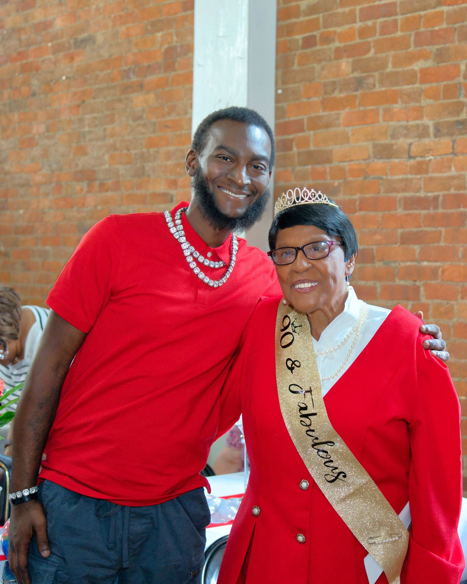 An older woman in a red shirt and a younger man in a red shirt are standing together and smiling.