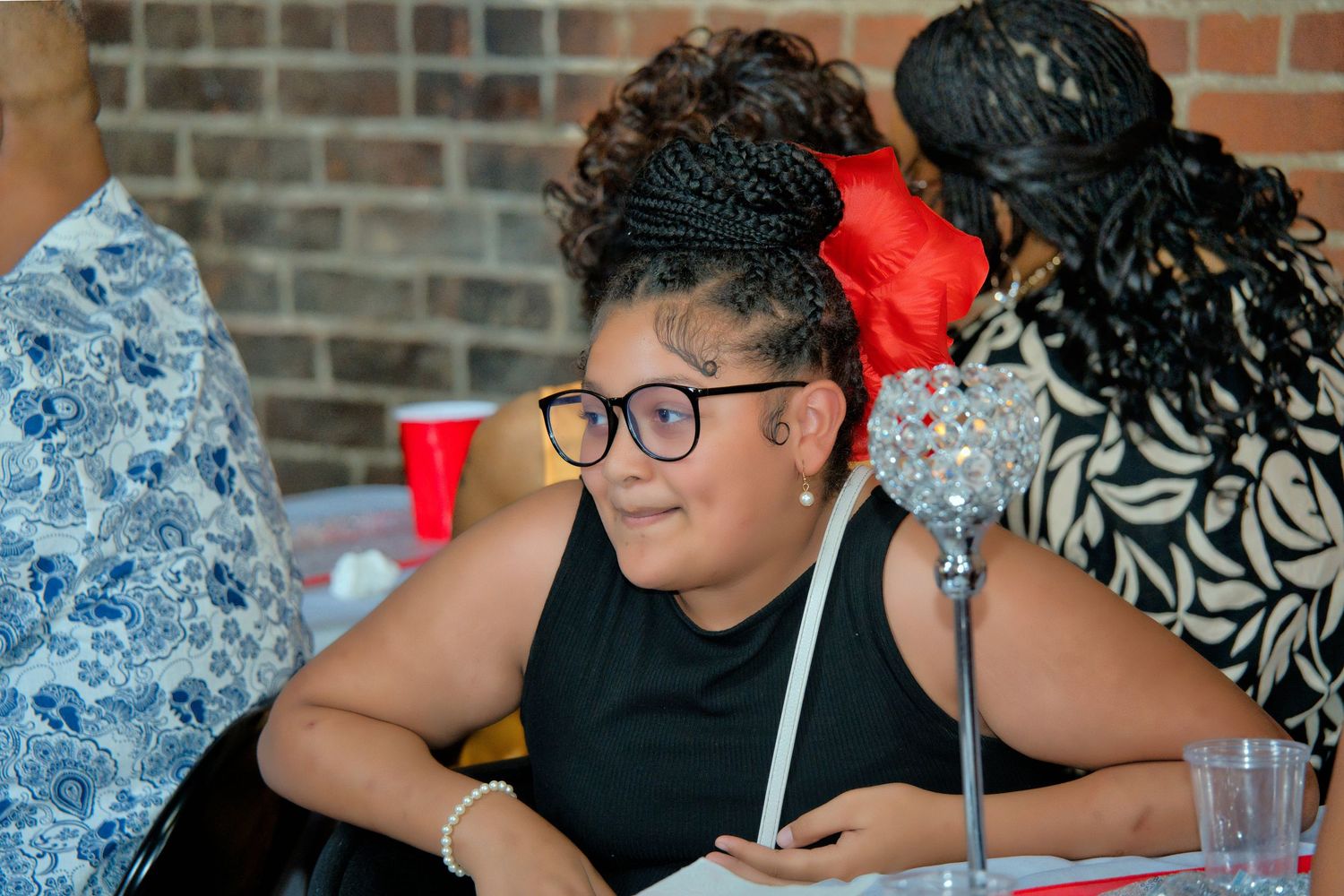A woman with curly hair and glasses is sitting at a table, smiling and looking at the camera.