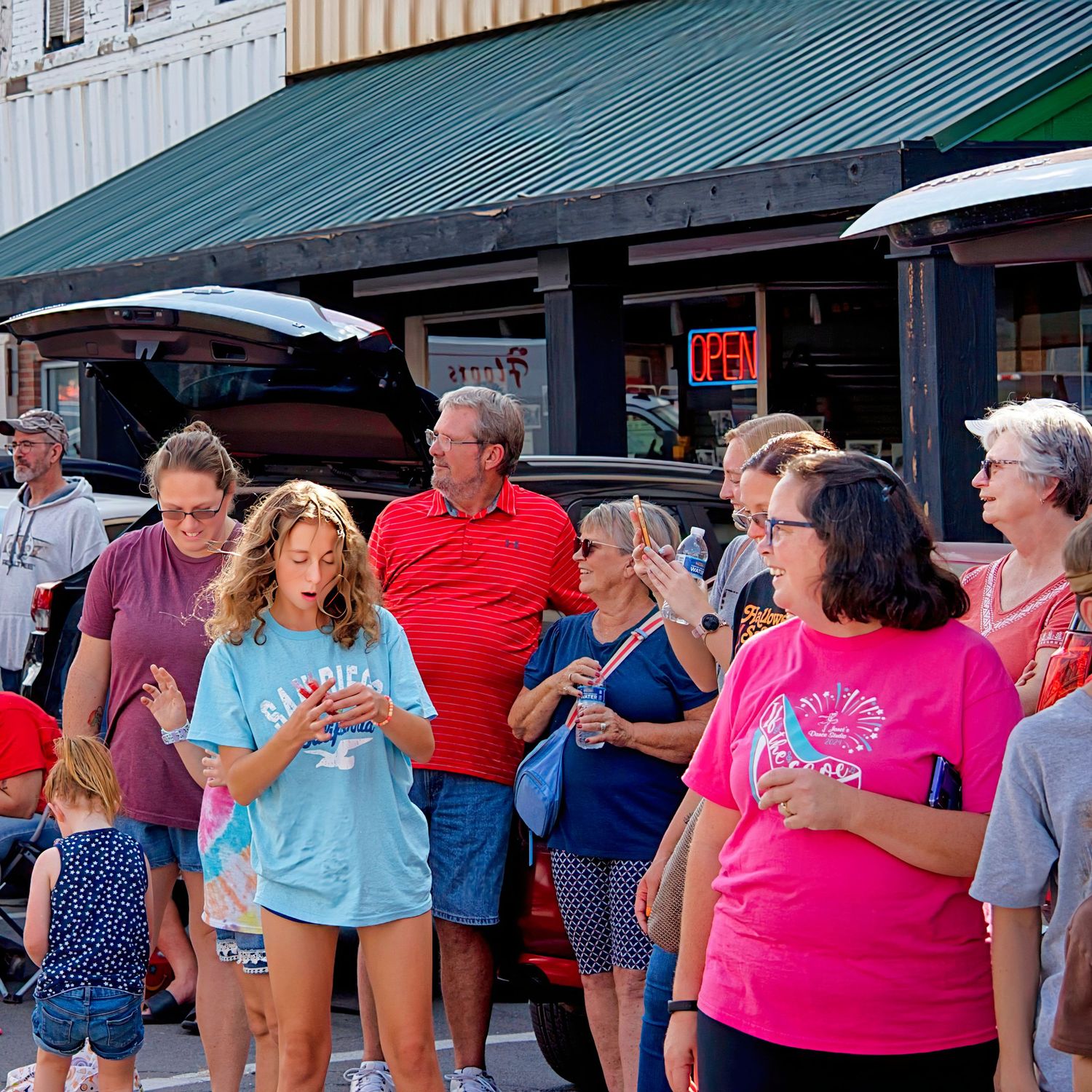 A crowd of people, some wearing colorful shirts, are gathered on a street with shops and buildings.