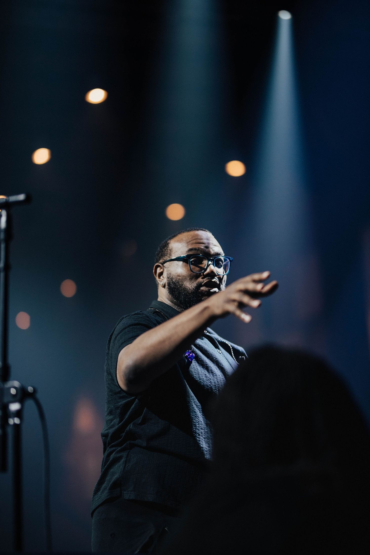 Black male performer gesturing on stage under dramatic blue and gold spotlights at live event.