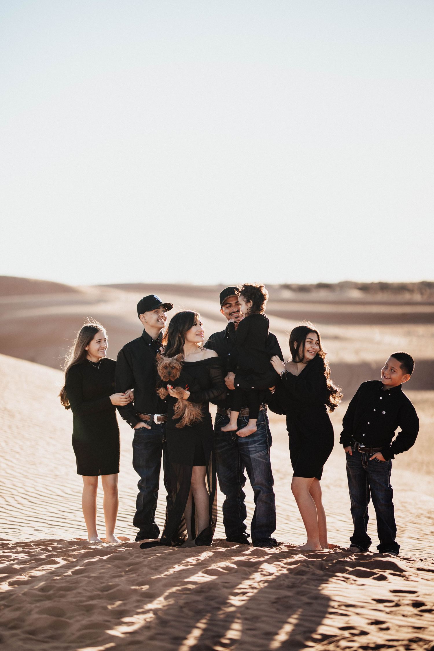 A large family of seven dressed in black poses together on sun-drenched sand dunes.