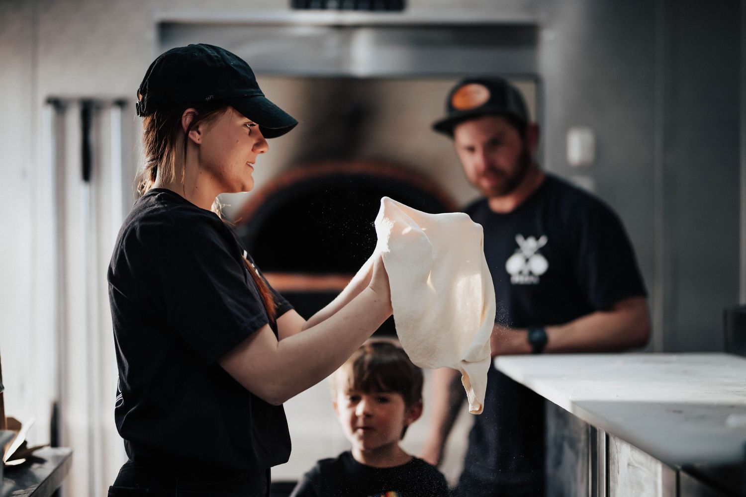 Pizza chef stretching dough by hand in a restaurant kitchen with a wood-fired oven in the background.