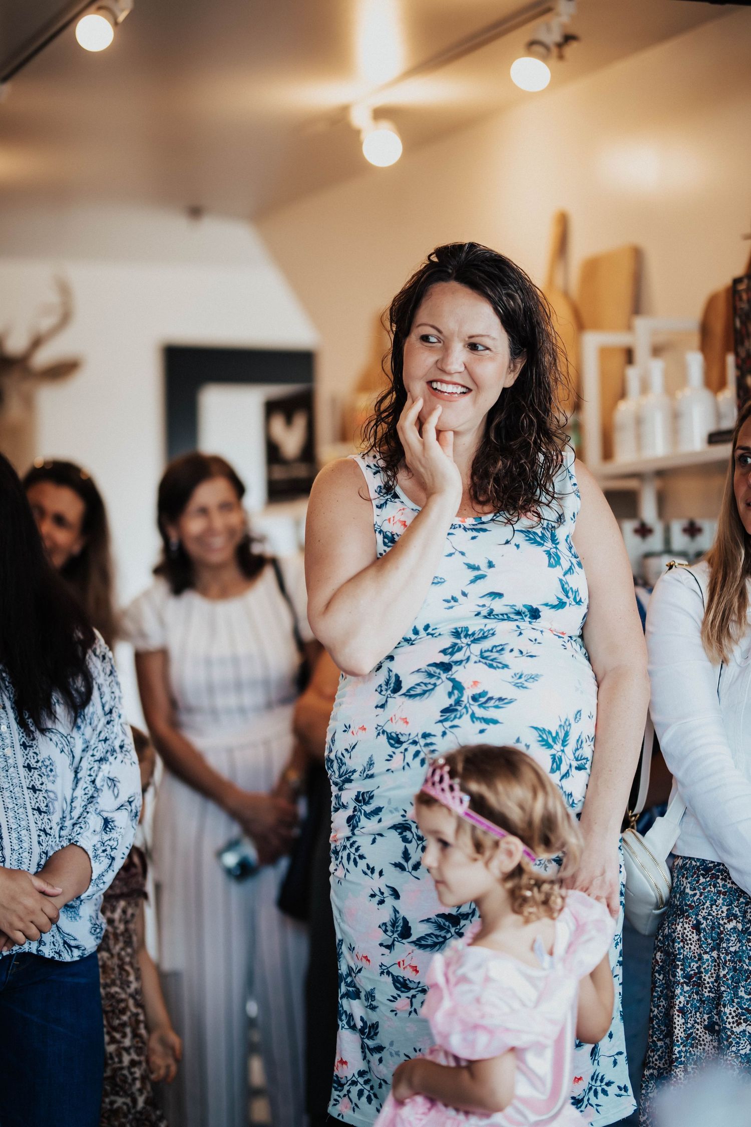 A smiling pregnant woman in a floral dress stands at her baby shower surrounded by guests and a young girl.