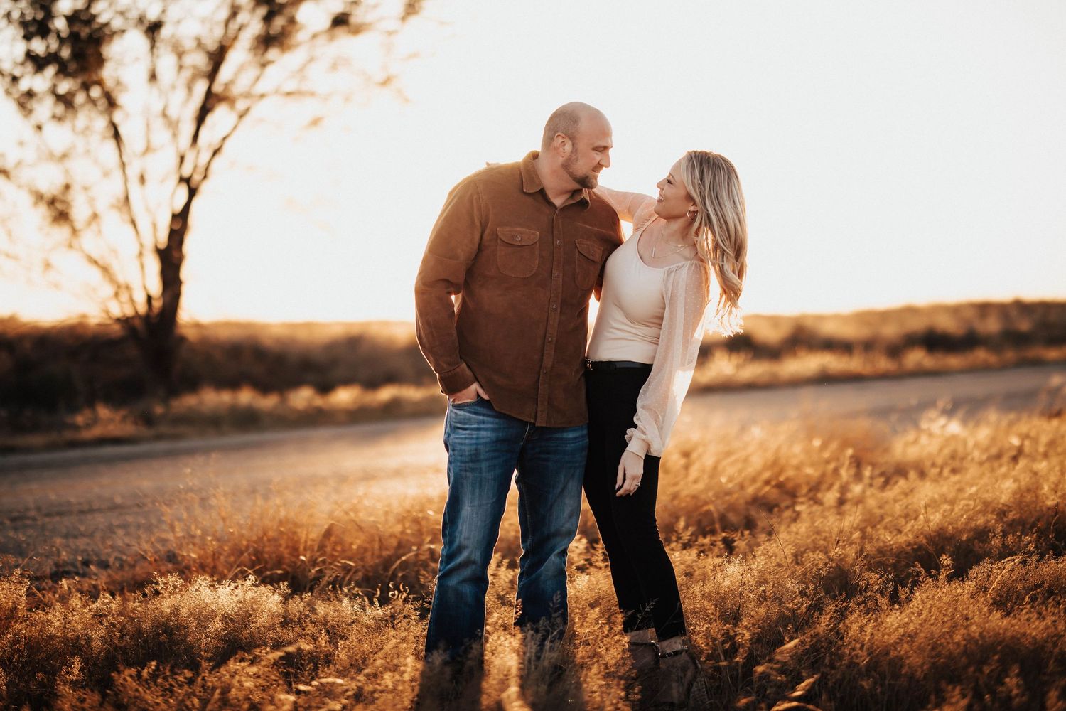 Couple standing close in golden sunset field, woman in white sweater leaning on mans shoulder.