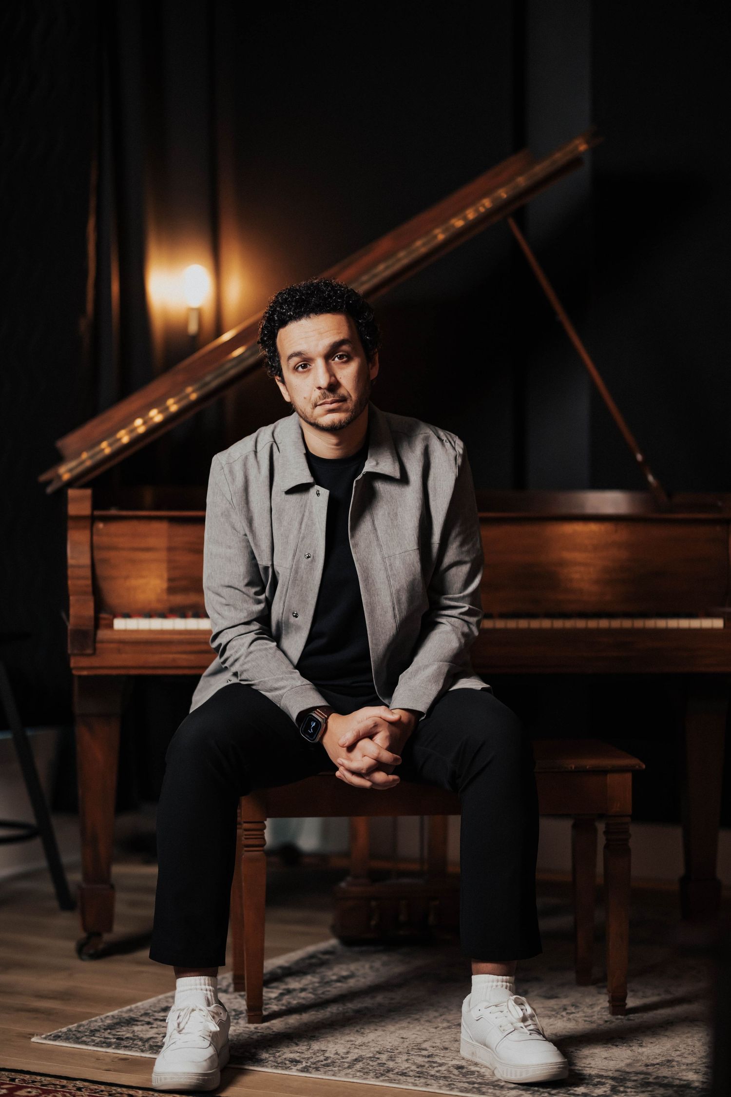 Man in gray jacket sits at a grand piano bench in a dimly lit studio, looking thoughtfully at the camera.