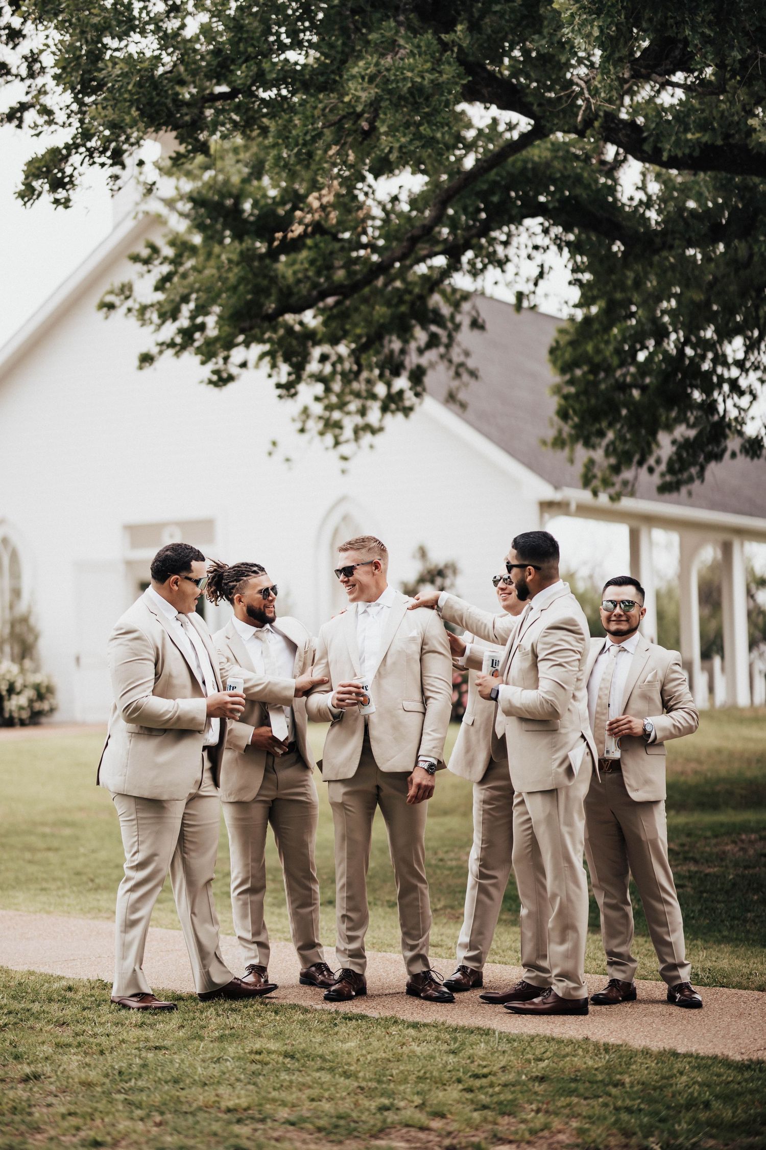 Groomsmen in matching tan suits and sunglasses gather outdoors near a white chapel on a wedding day.