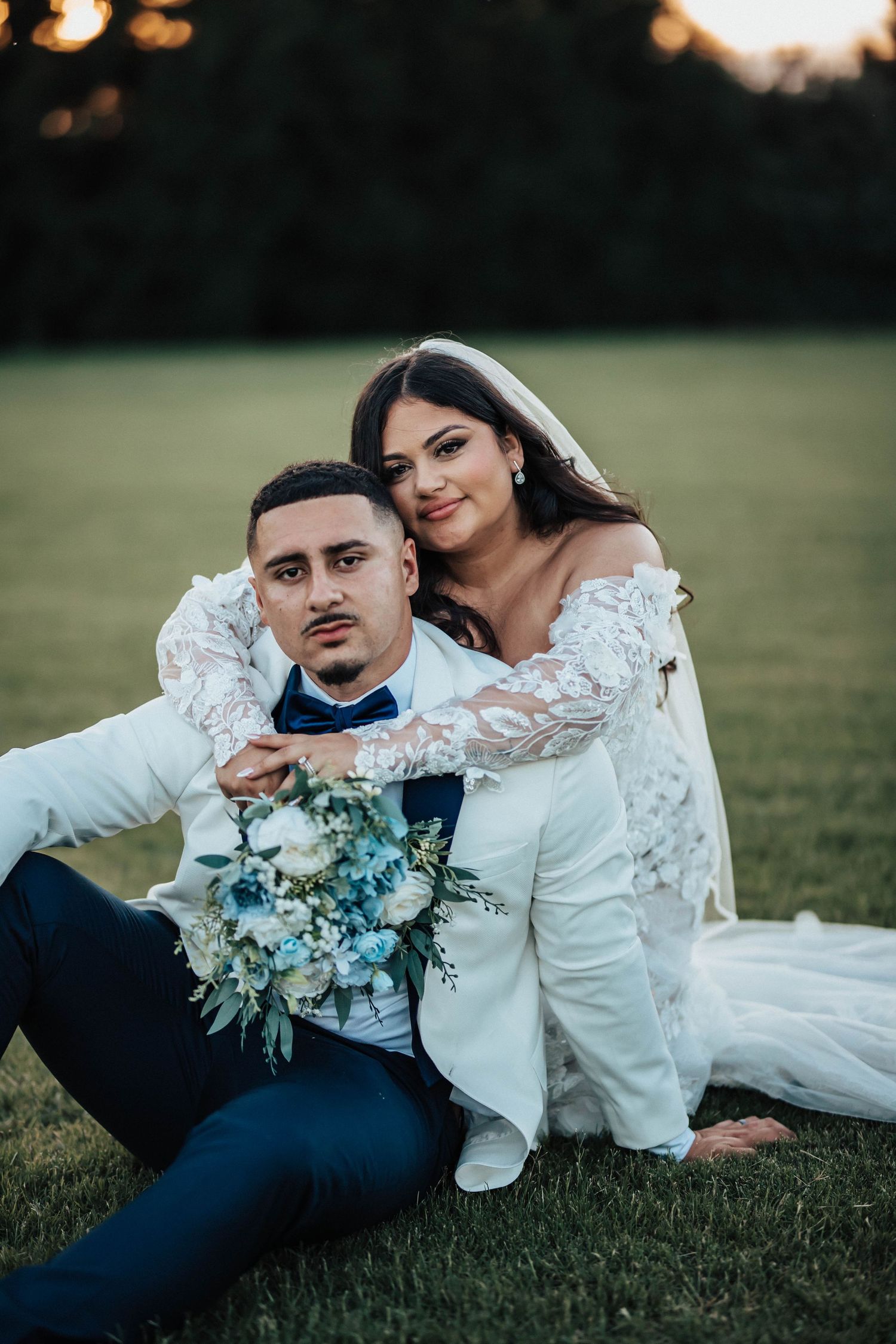 Bride and groom sitting on green grass, bride in lace off-shoulder gown holding blue and white bouquet, groom in white suit.