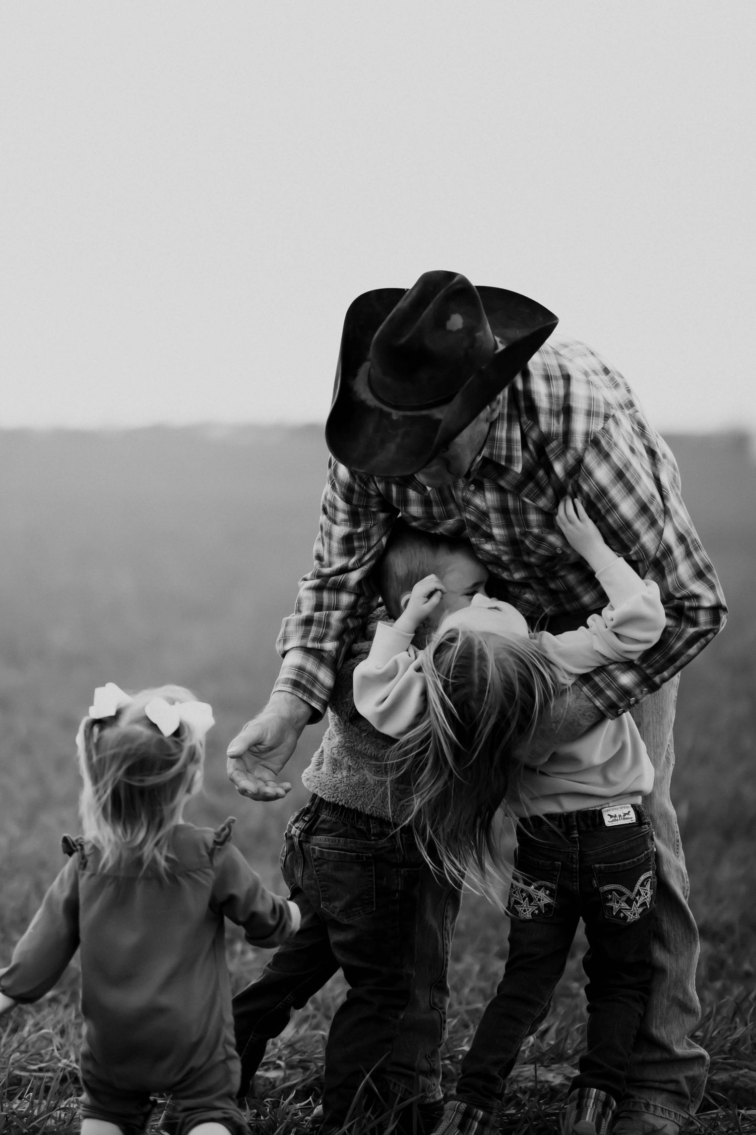 Black and white photo of father in cowboy hat hugging young children in field.