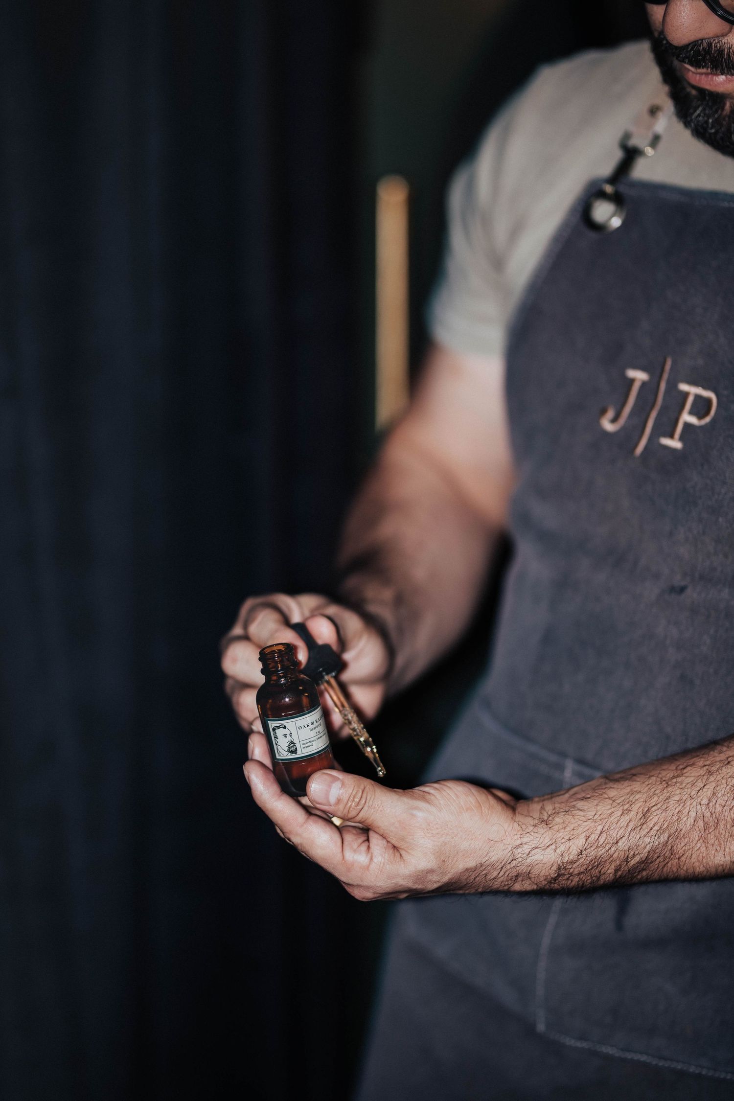 Barber in denim apron holding a small bottle of beard oil in a barbershop.
