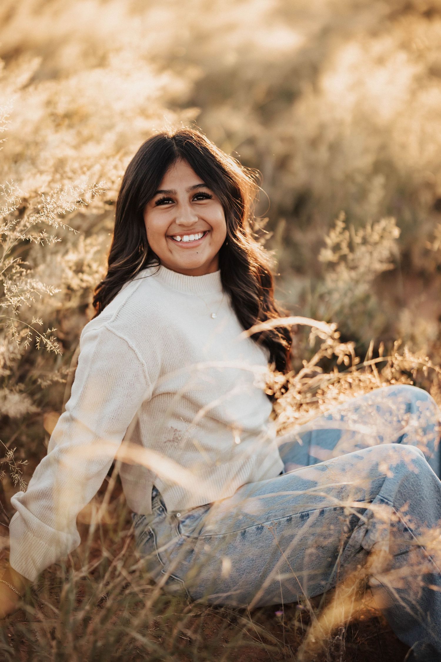 Young woman with dark hair smiling in a golden field, wearing a white sweater and jeans during golden hour.