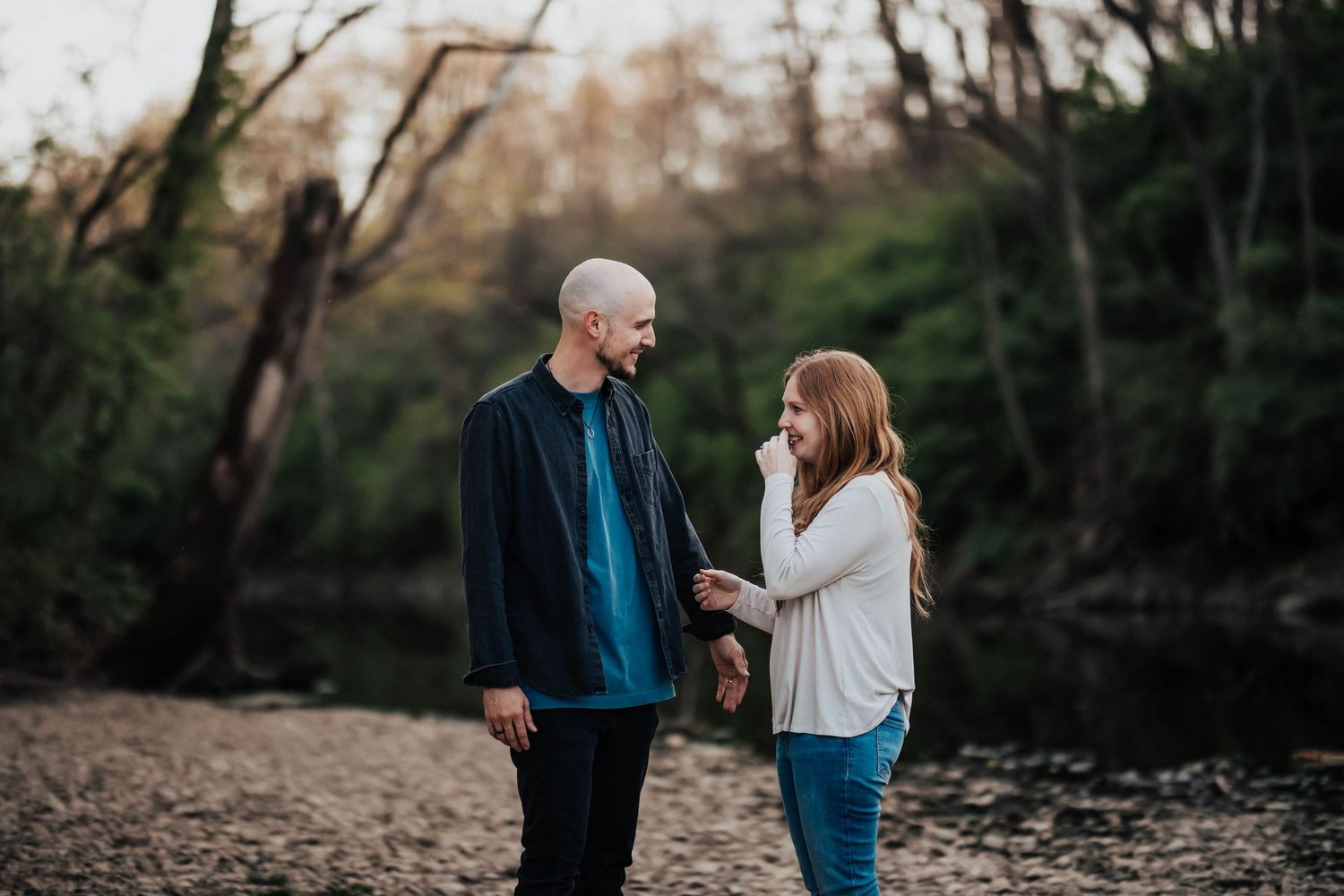 Couple holding hands by a forest creek, woman with red hair smiling at bald man in blue jacket.