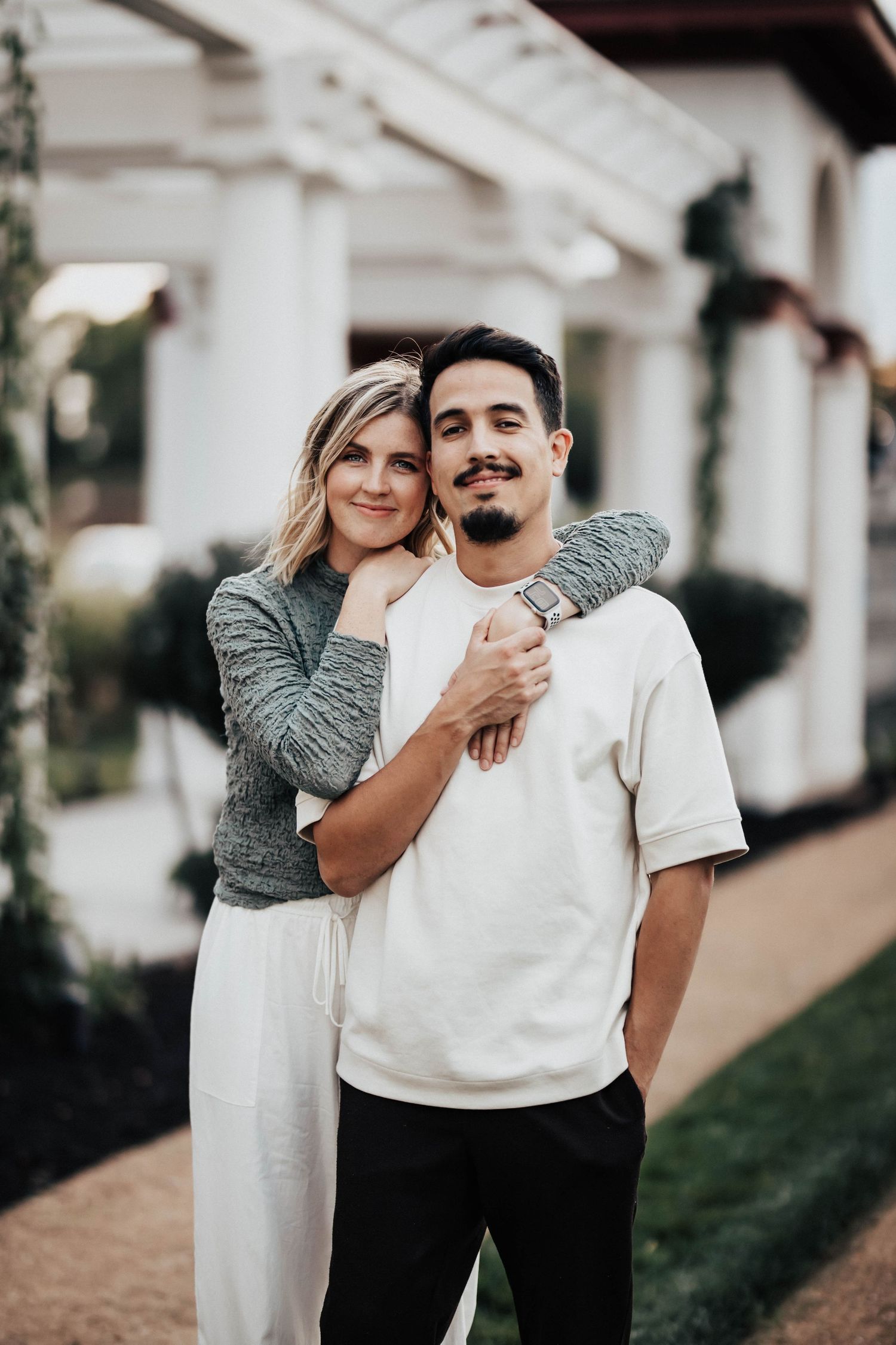 Couple posing together outdoors, woman in gray sweater hugging man in white shirt from behind.