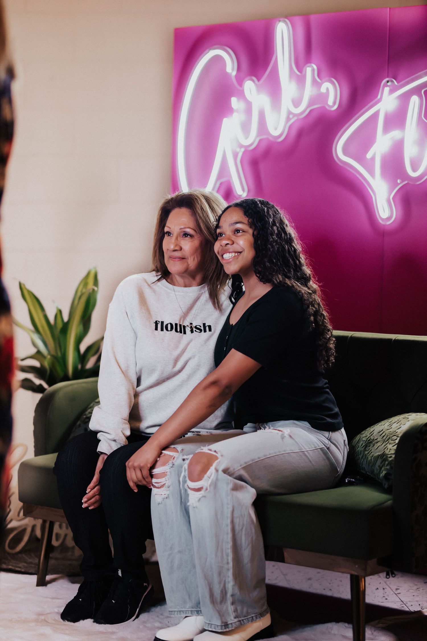 Two women pose together smiling in front of a pink Girls Flourish neon sign.