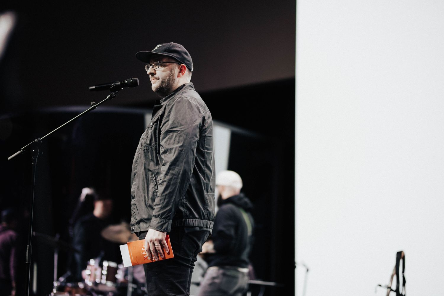 Man in cap and jacket speaks at microphone on stage with band members in background.