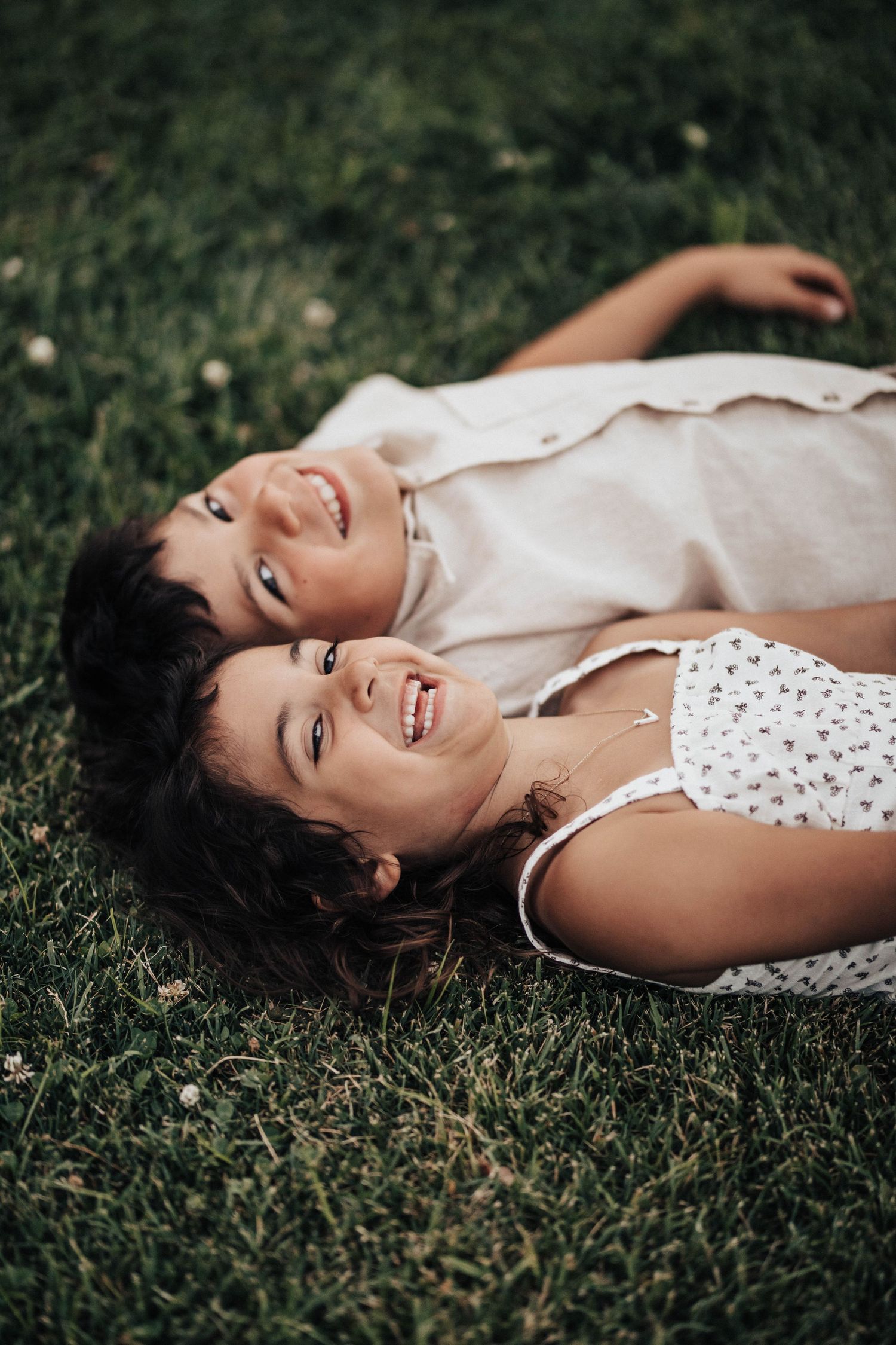 Two smiling children lying on green grass together, enjoying a sunny outdoor day.