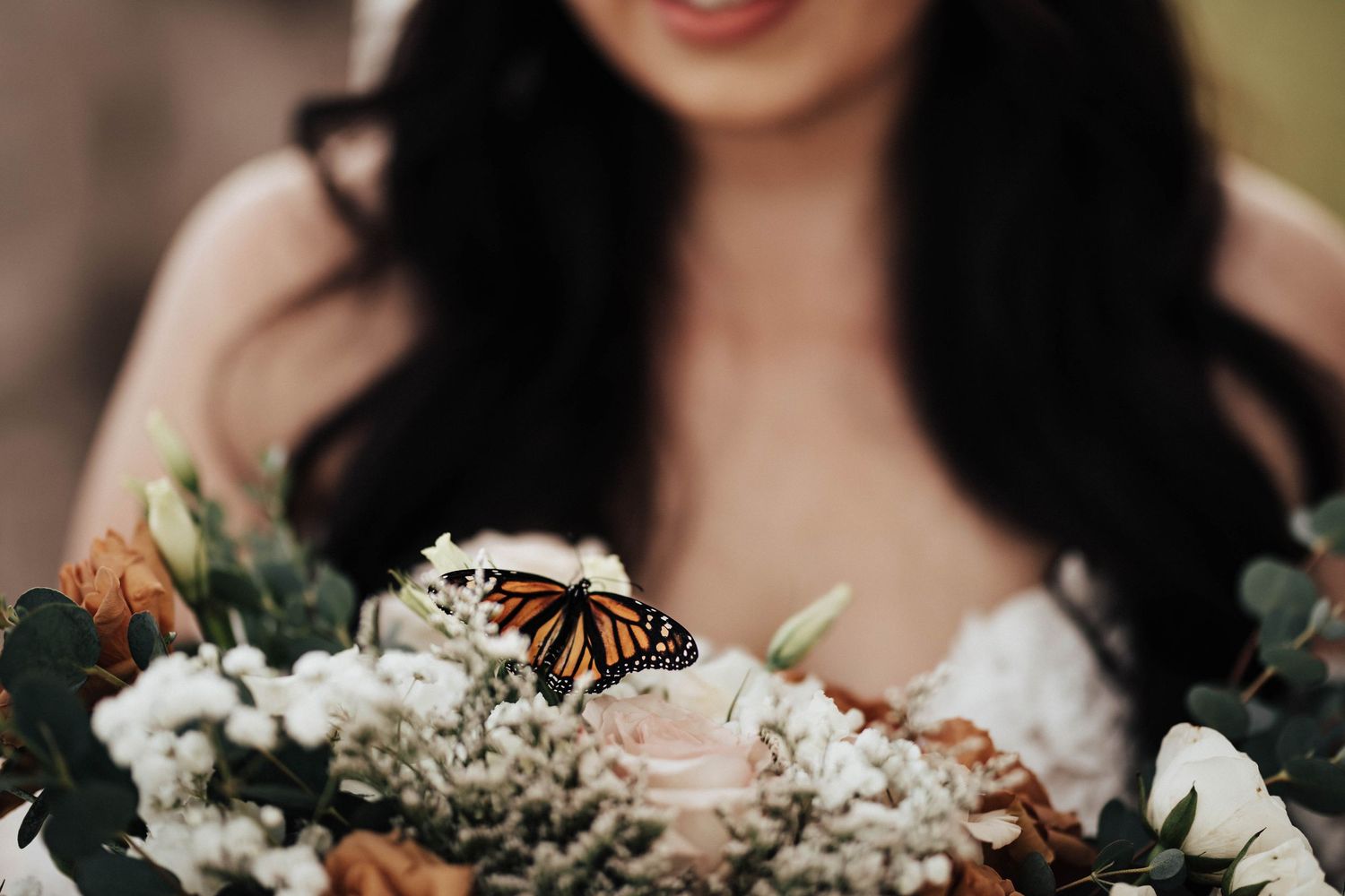 A monarch butterfly perches on a bridal bouquet of white and orange flowers held by a dark-haired bride.