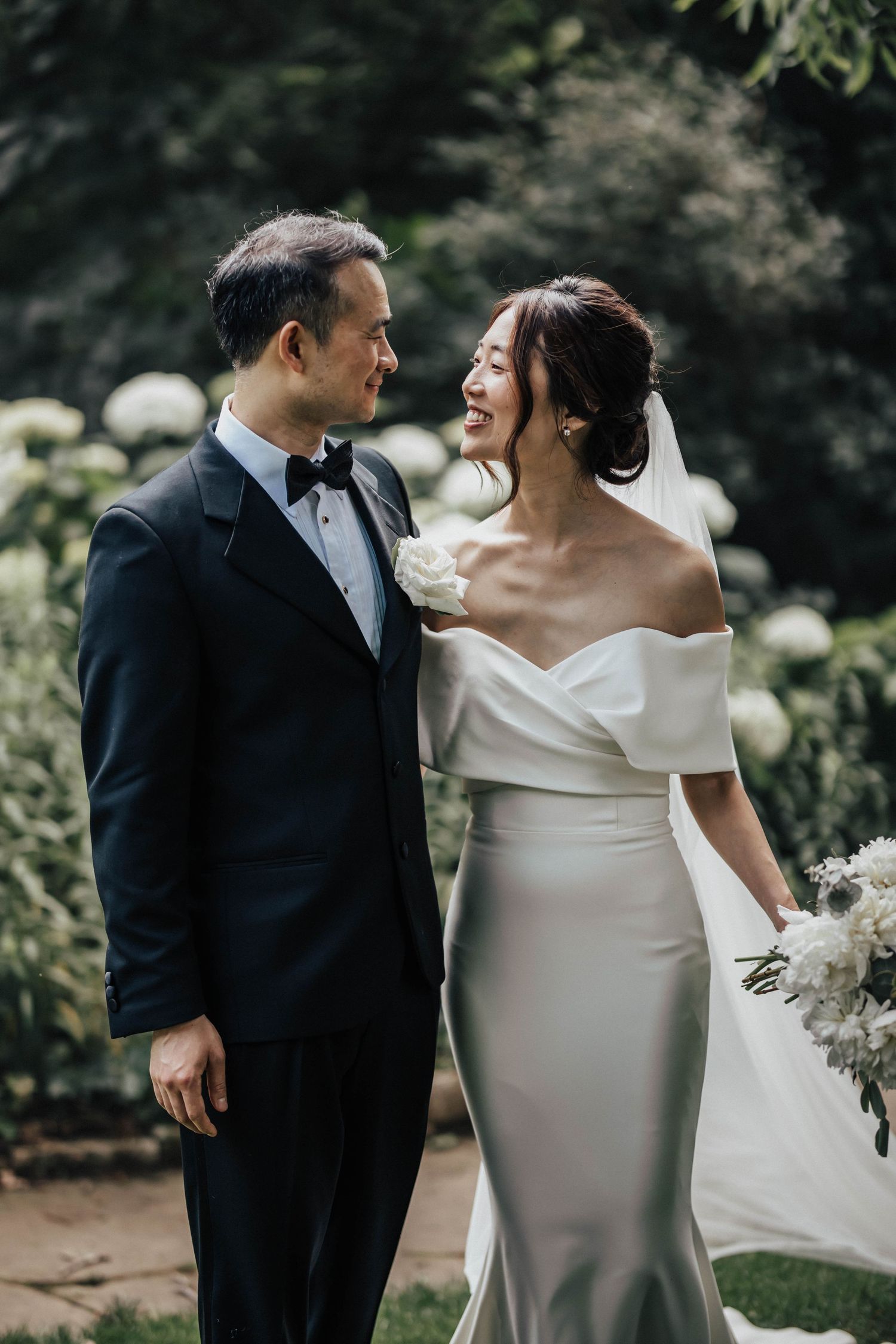 A bride and groom share a joyful moment in a garden, she in an off-shoulder white gown, he in a classic black tuxedo.