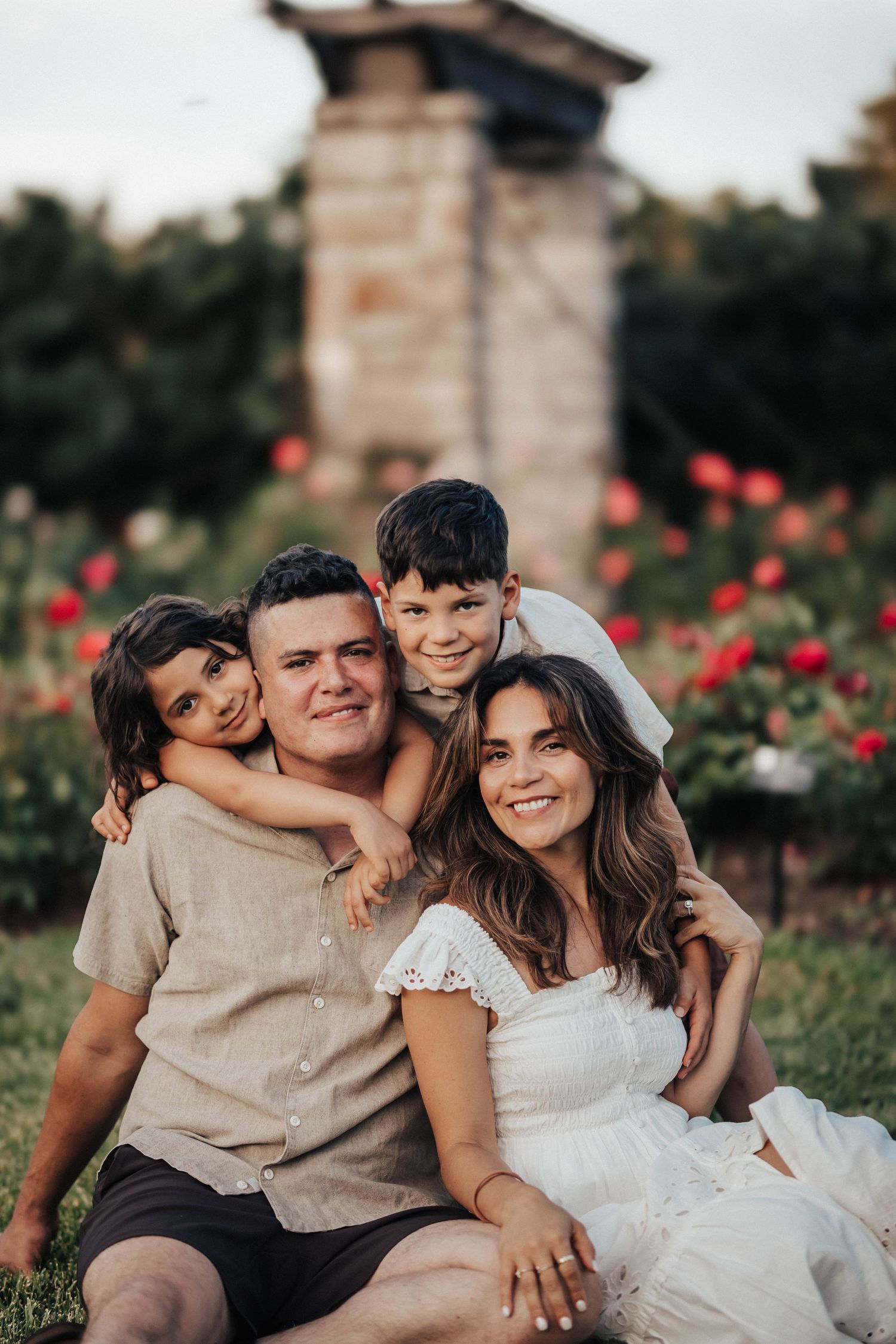 Happy family of four posing outdoors in a rose garden, parents smiling with two young children hugging them.