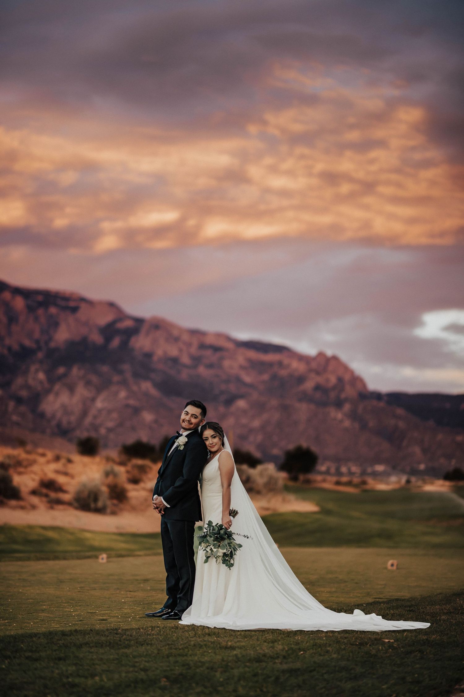 Bride and groom posing at sunset with dramatic mountain backdrop on golf course.