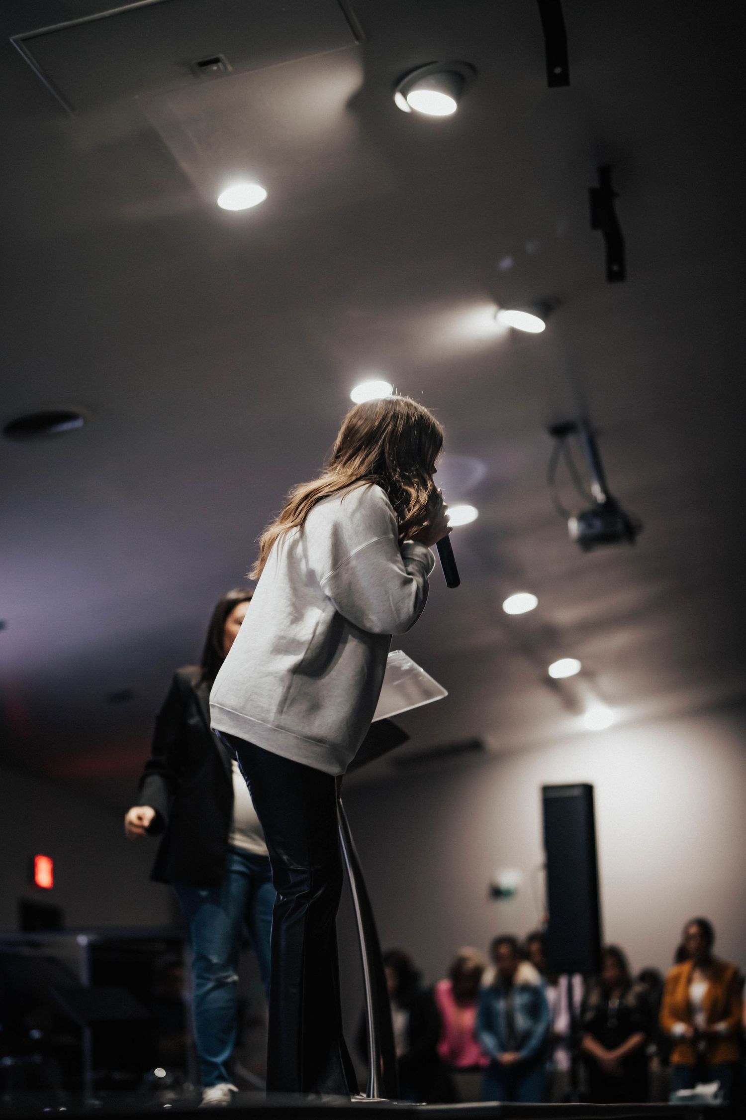 A woman speaks into a microphone on stage at a church event with audience seated below.