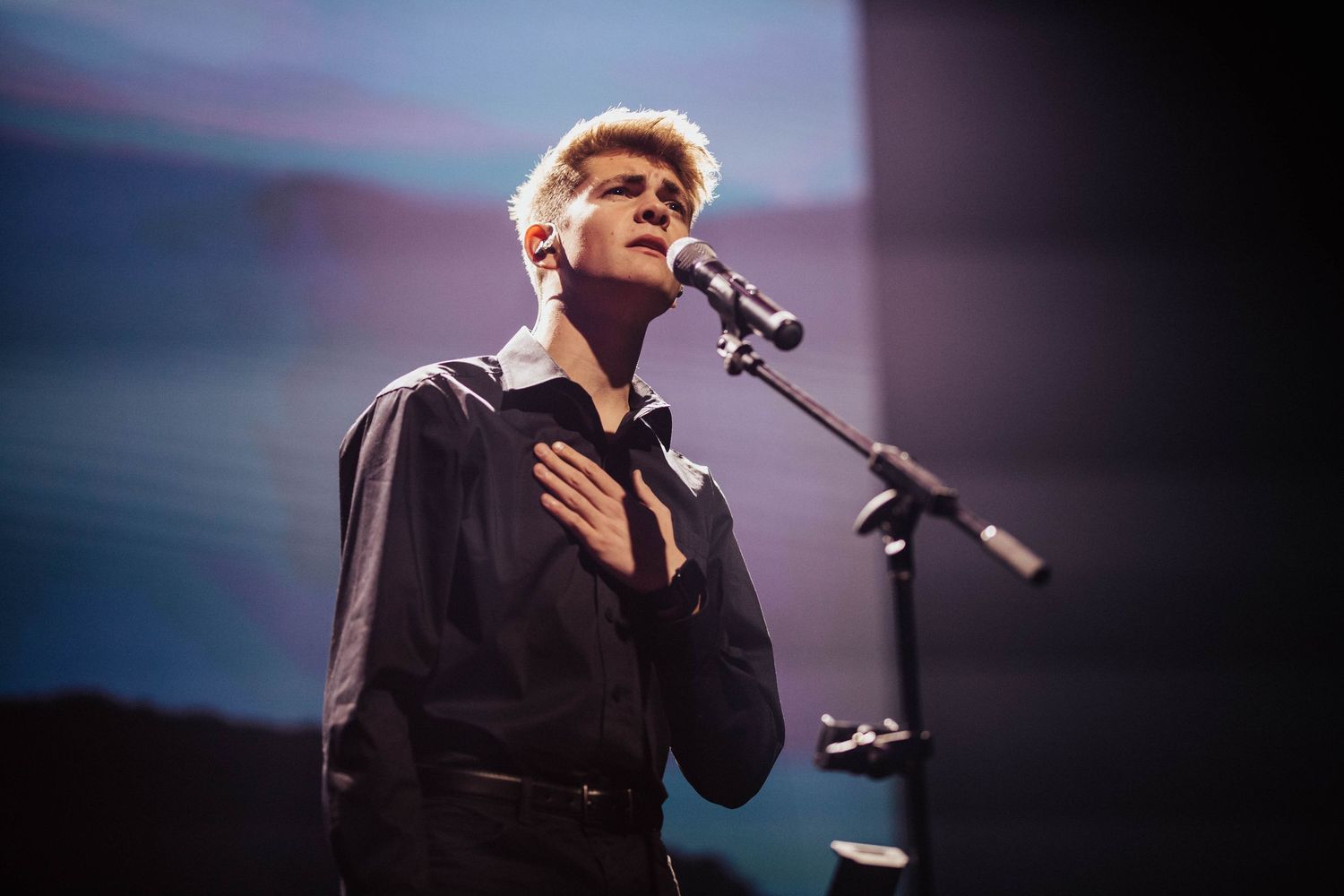 A young male singer performs on stage at a microphone, hand on chest, under dramatic blue stage lighting.