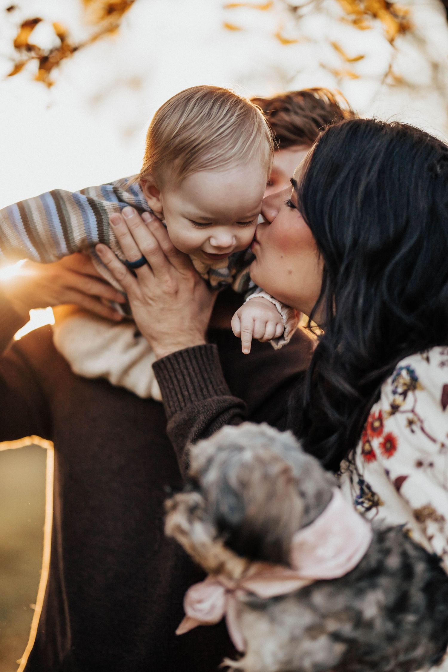 Mother kissing smiling baby cheek while father holds child in a warm golden light.