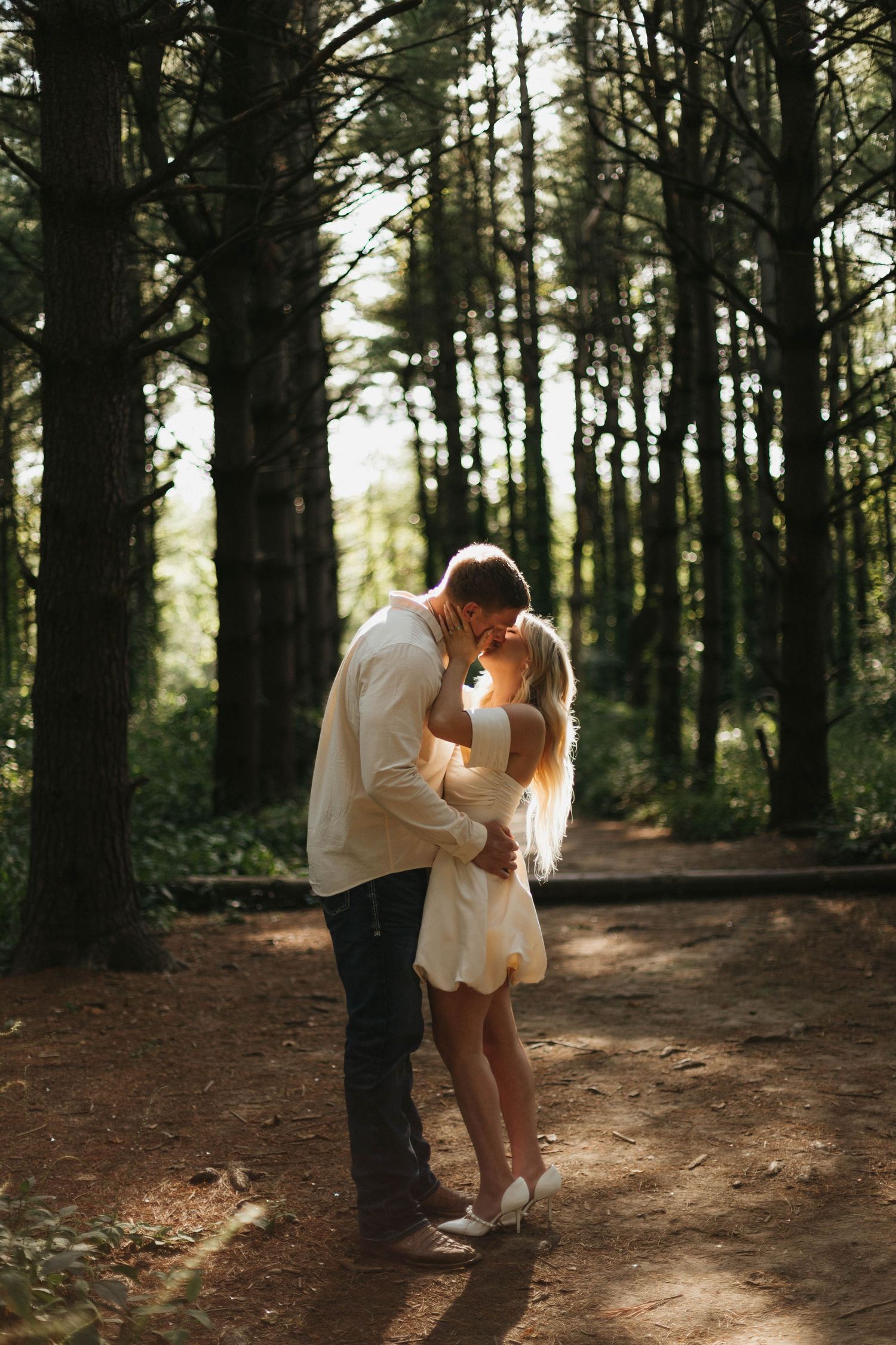 Couple sharing a romantic kiss on a sunlit forest path surrounded by tall trees in a golden hour outdoor setting.