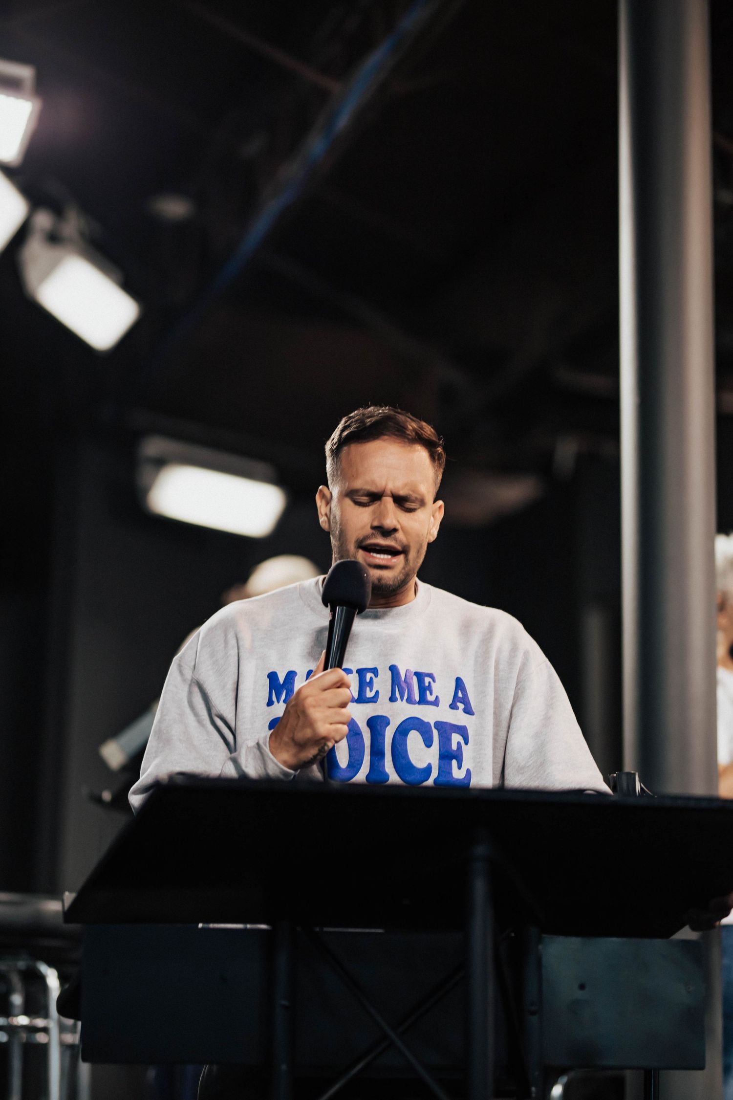 A man in a Make Me a Voice sweatshirt leads worship at a podium with a microphone.