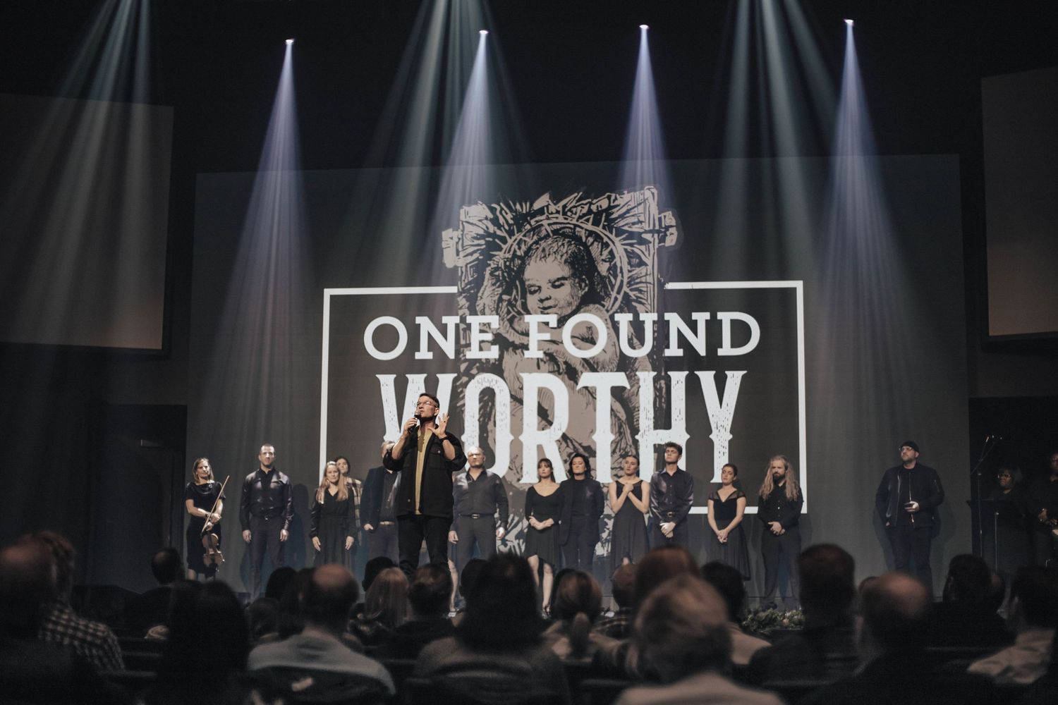 A large ensemble cast stands on a lit stage beneath an One Found Worthy sign during a live performance.