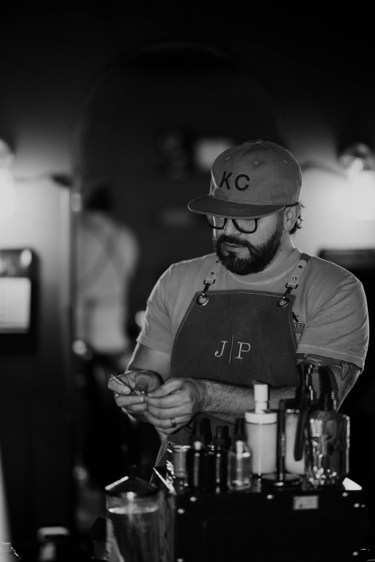 Bearded barber in KC cap and denim apron working at a barbershop counter in black and white.