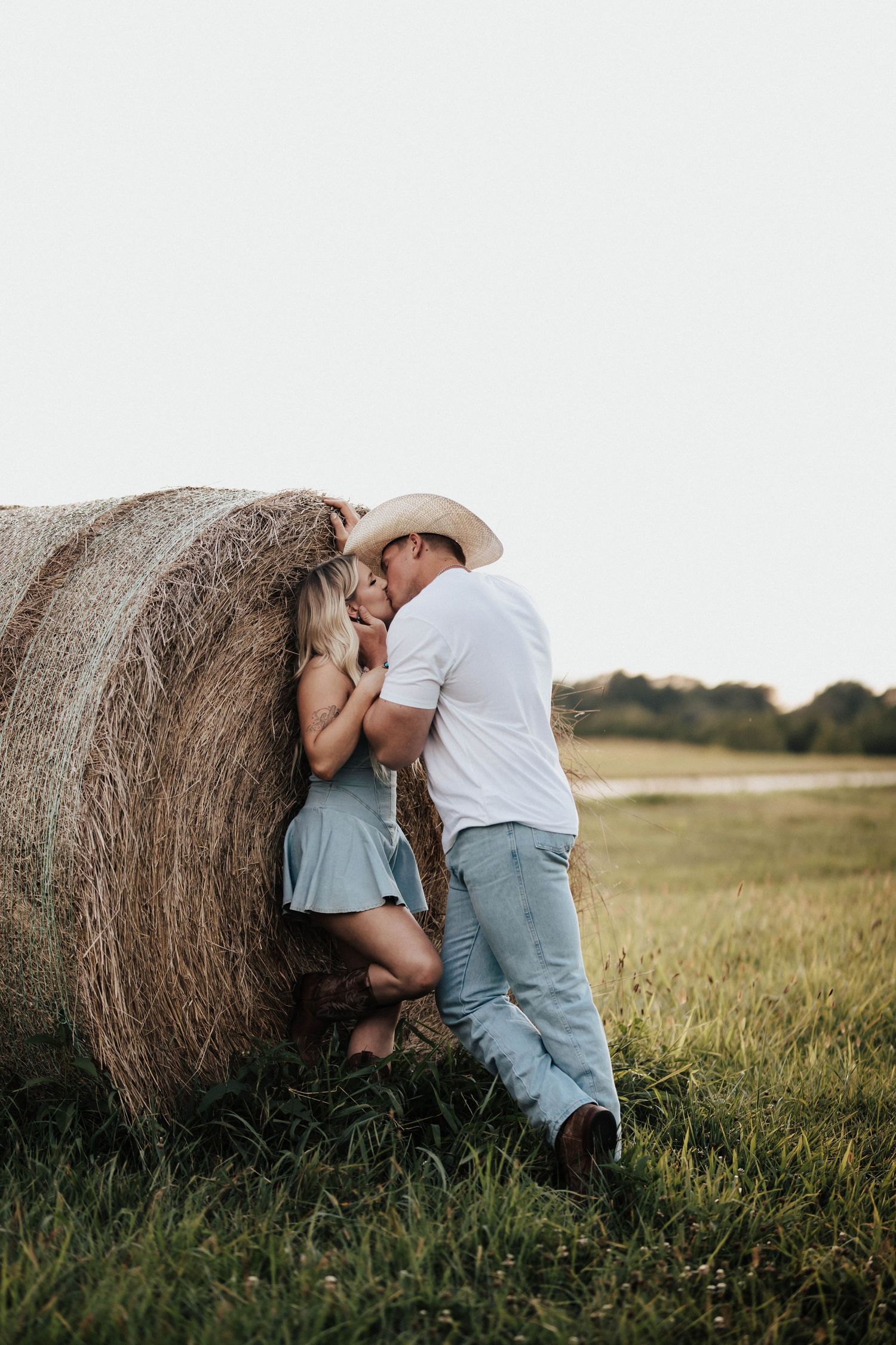 Couple sharing a romantic kiss leaning against a hay bale in a green field, man wearing cowboy hat and white shirt.