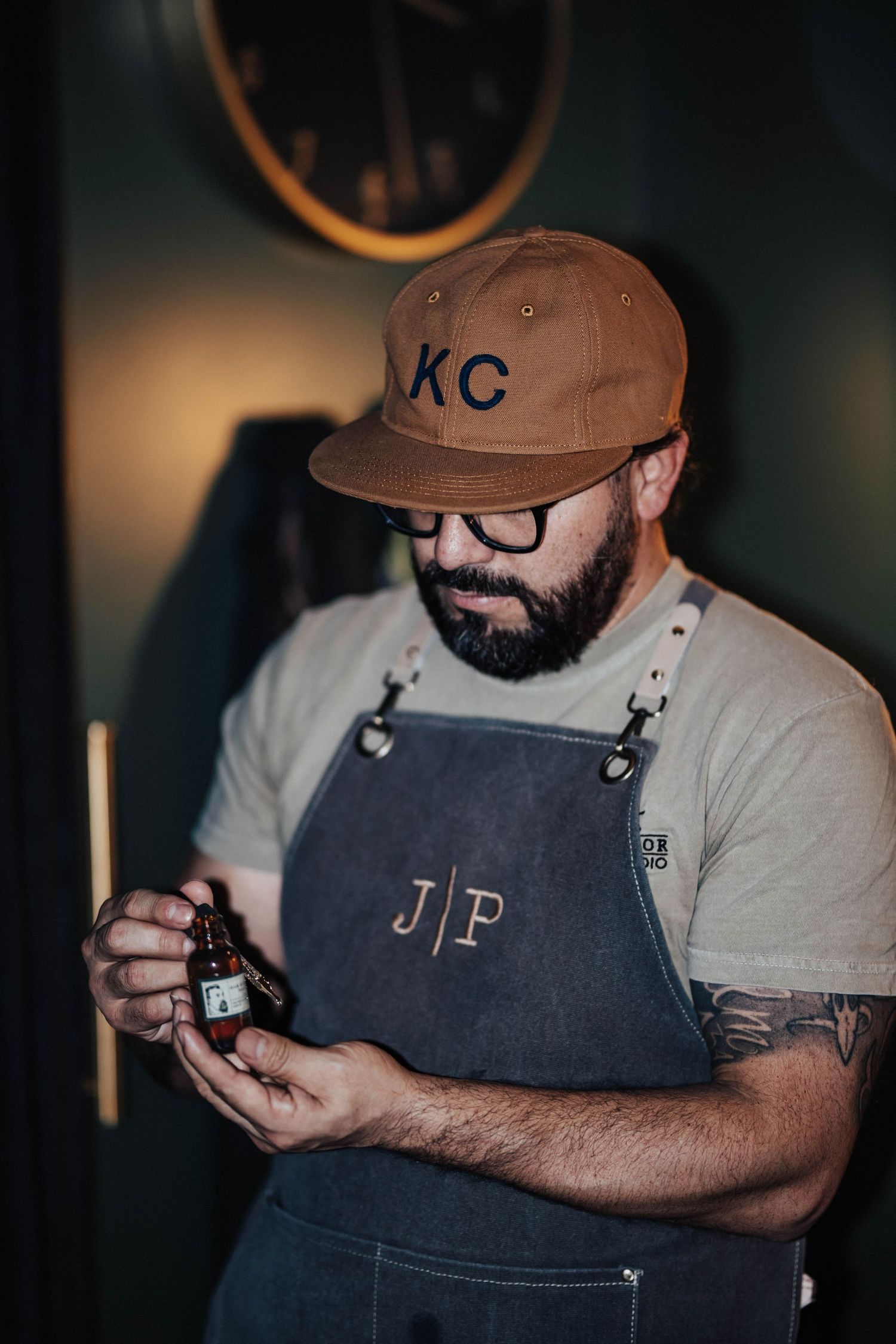 Bearded barber wearing a KC cap and denim apron examining a bottle of beard oil.