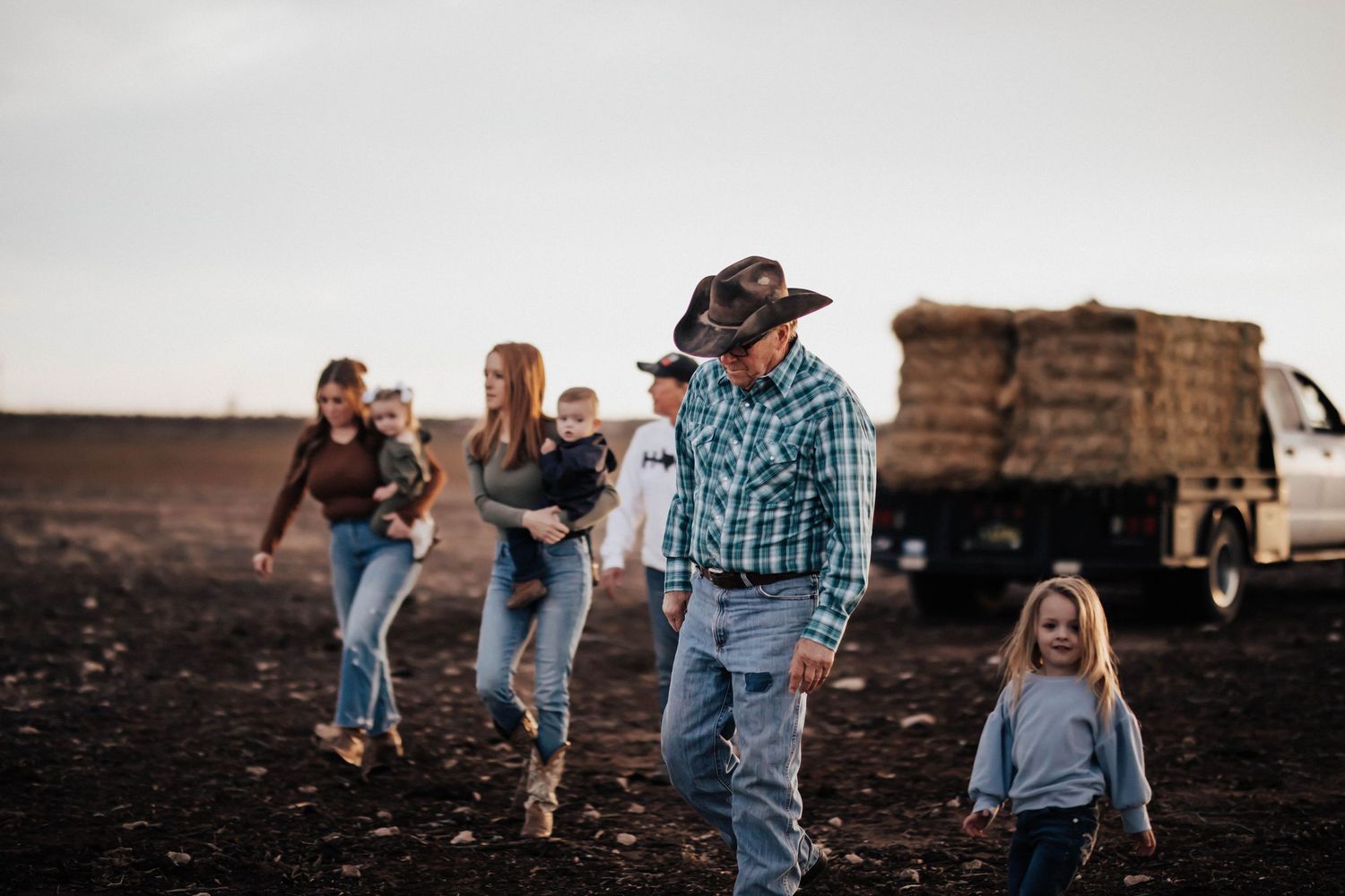 Family walking across farm field with cowboy hat dad leading young children.