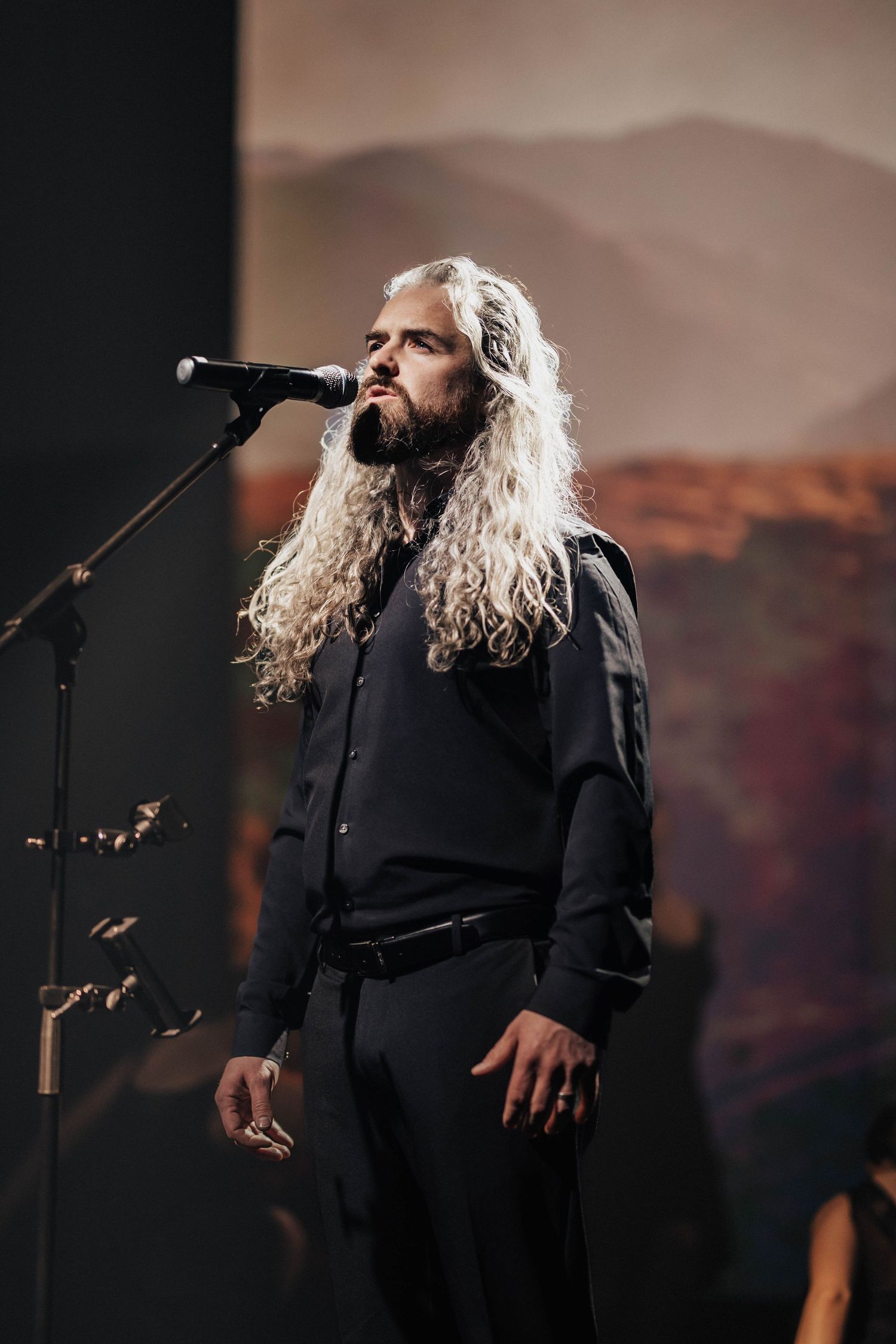 Long white-haired male singer performing at microphone on stage with scenic backdrop lighting.