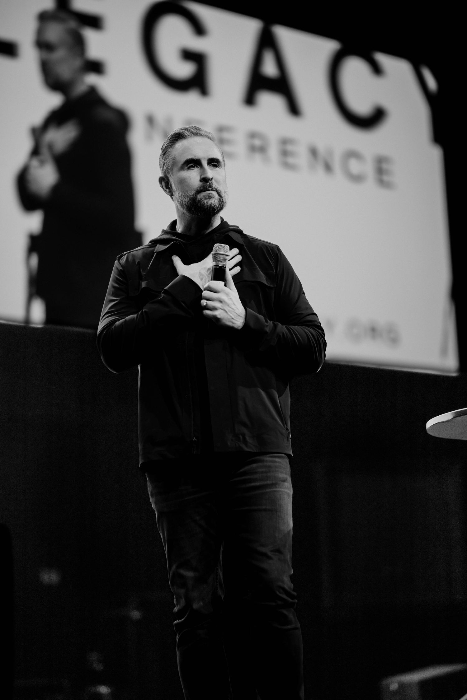 Black and white photo of a bearded man speaking on stage at the Legacy Conference, holding a microphone.