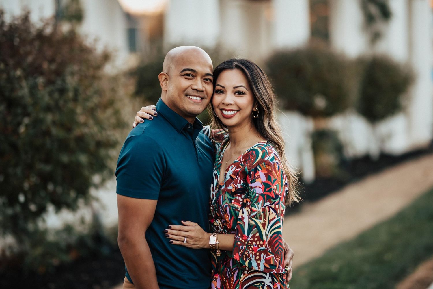 Smiling couple posing outdoors, man in blue polo with arm around woman in colorful floral dress.