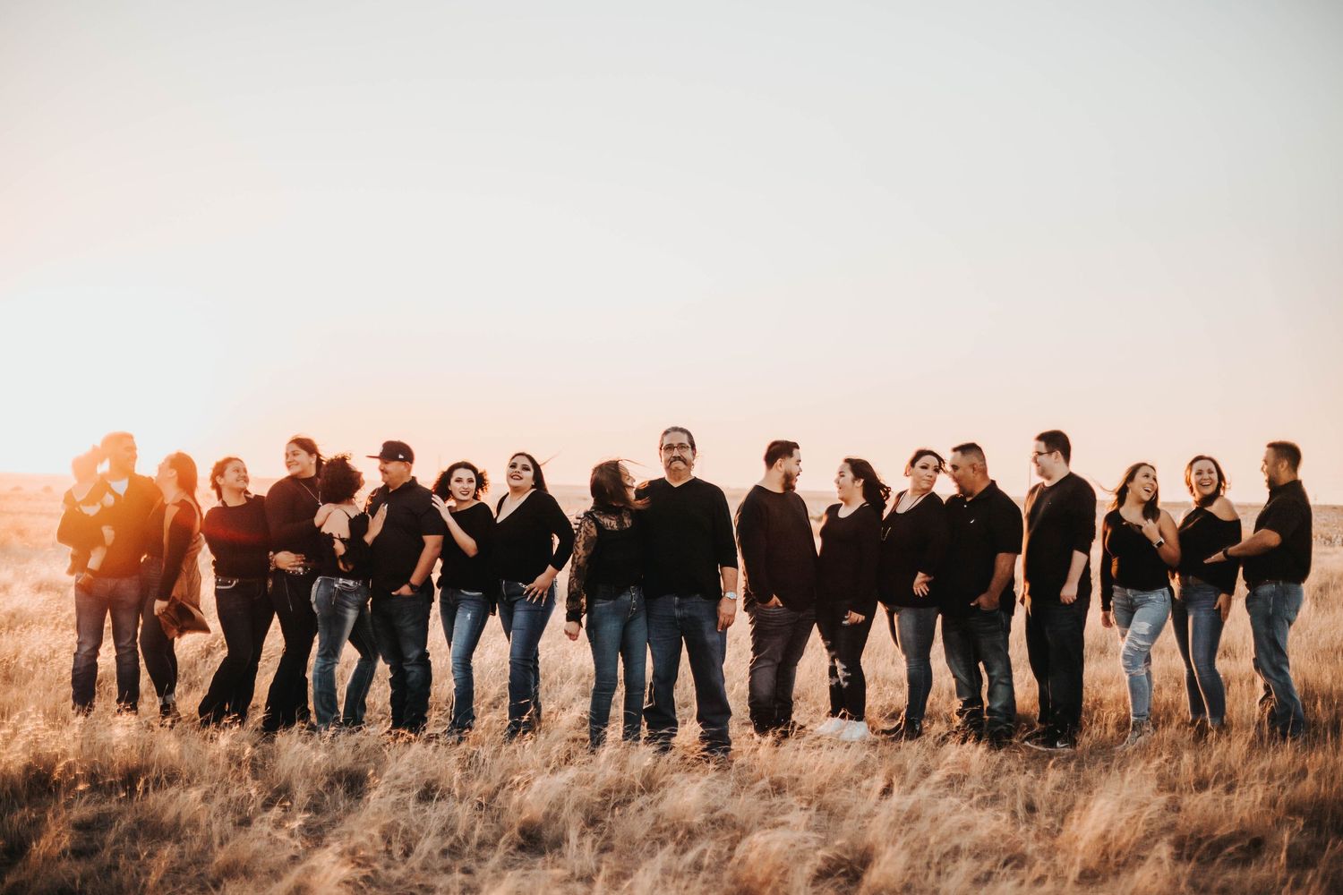 Large extended family group portrait in matching black outfits standing in golden field at sunset.