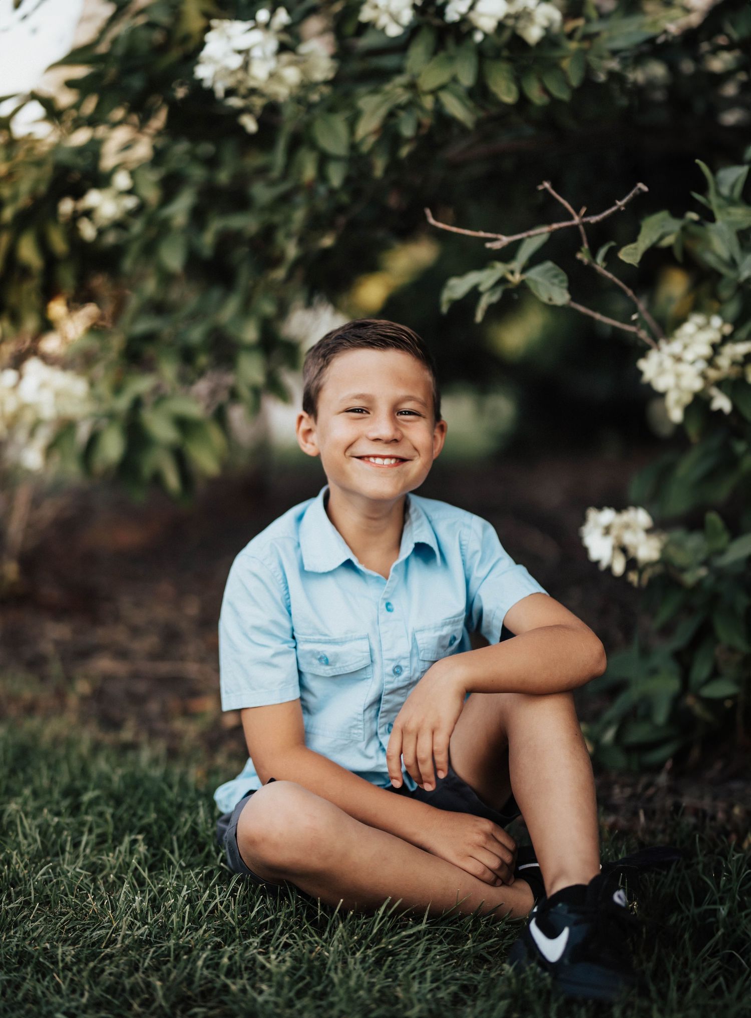 Smiling boy in light blue shirt sitting cross-legged on grass under flowering white blooms in a garden setting.